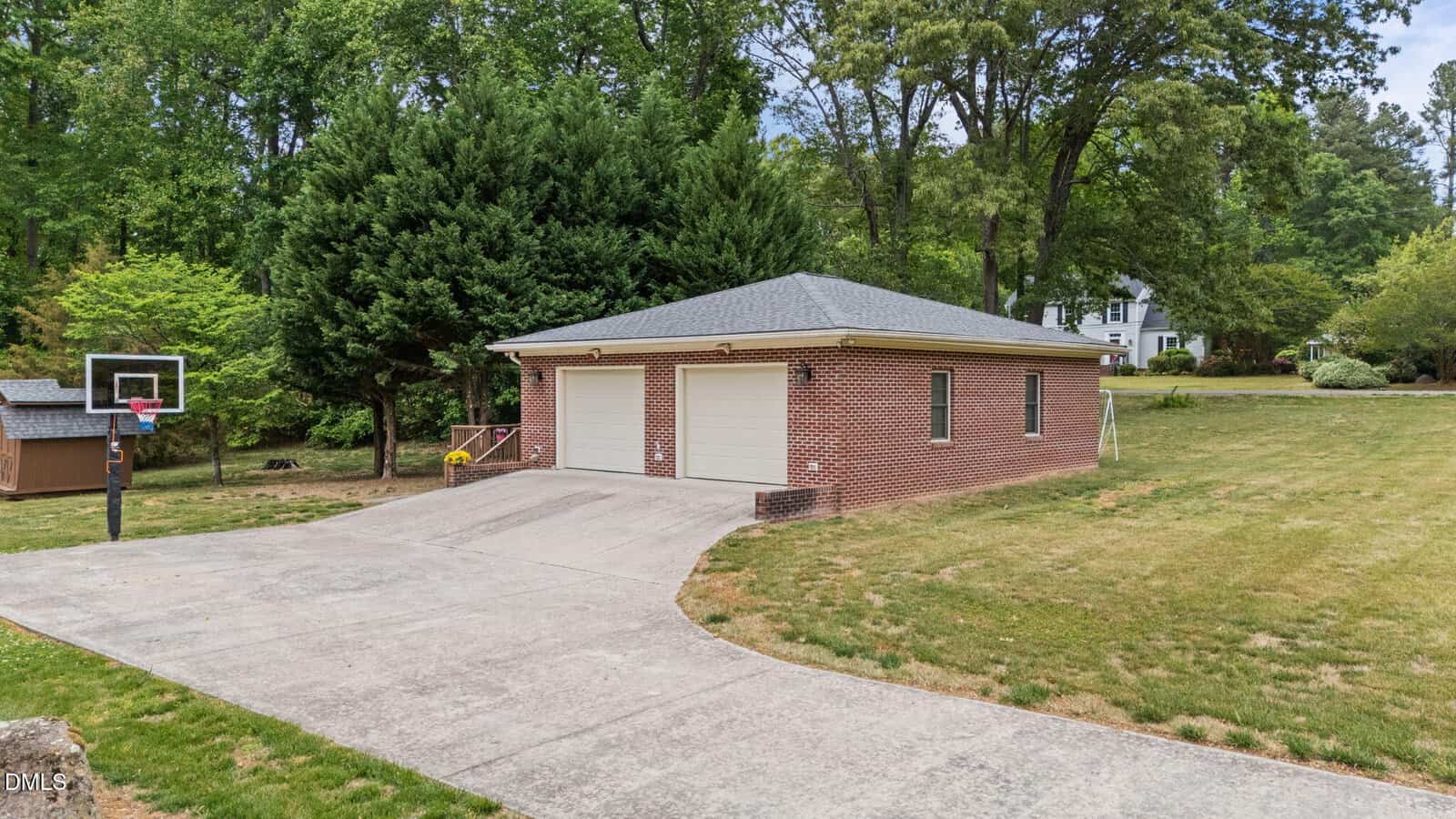 A brick two-car garage with a gray roof sits at the end of a wide concrete driveway, surrounded by green grass, trees, and a basketball hoop on the left. A house is visible in the background among the trees.