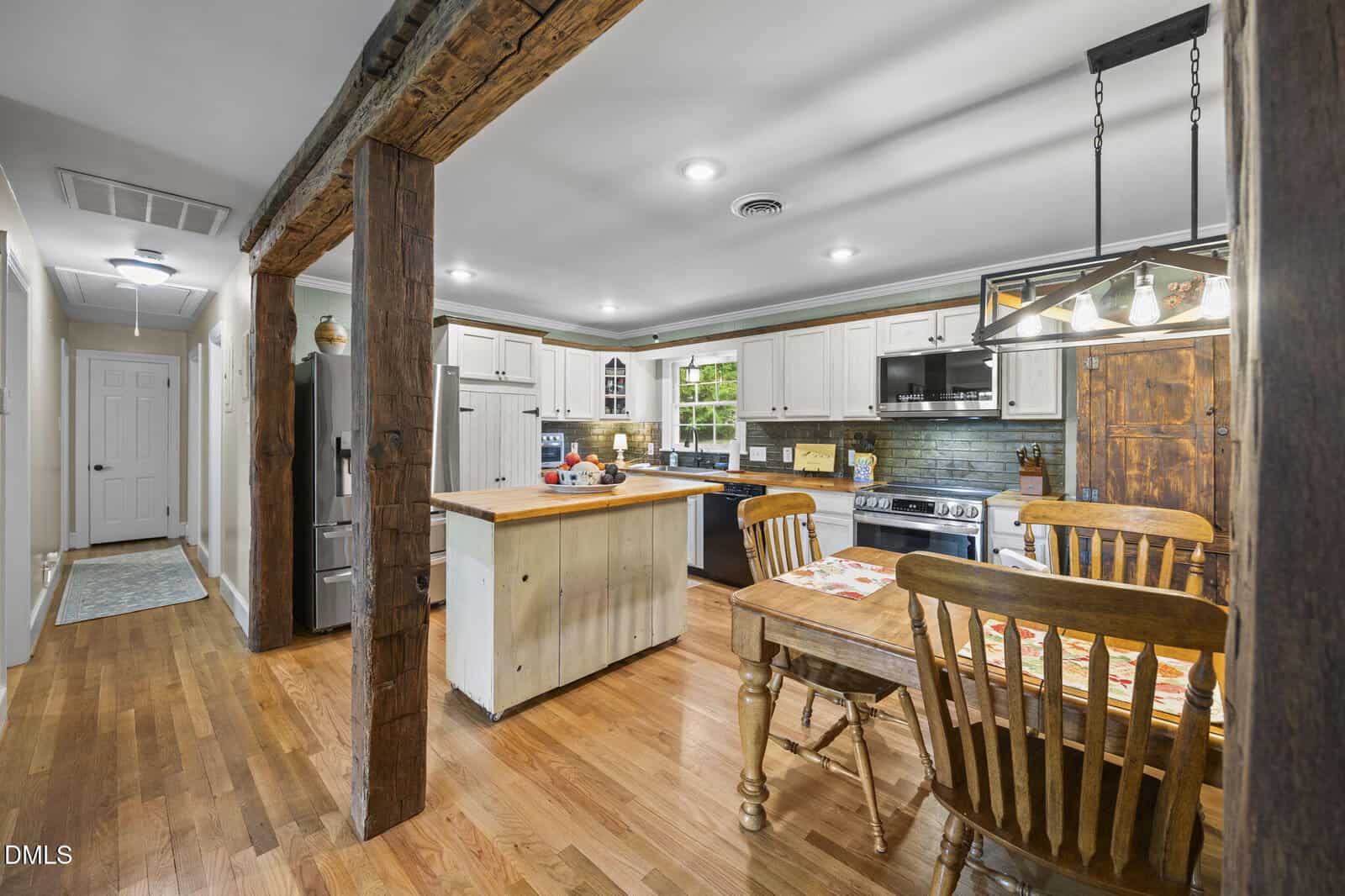 A rustic kitchen with wooden floors, exposed beams, white cabinets, a central island, a dining table with four chairs, stainless steel appliances, and a modern light fixture above the table.