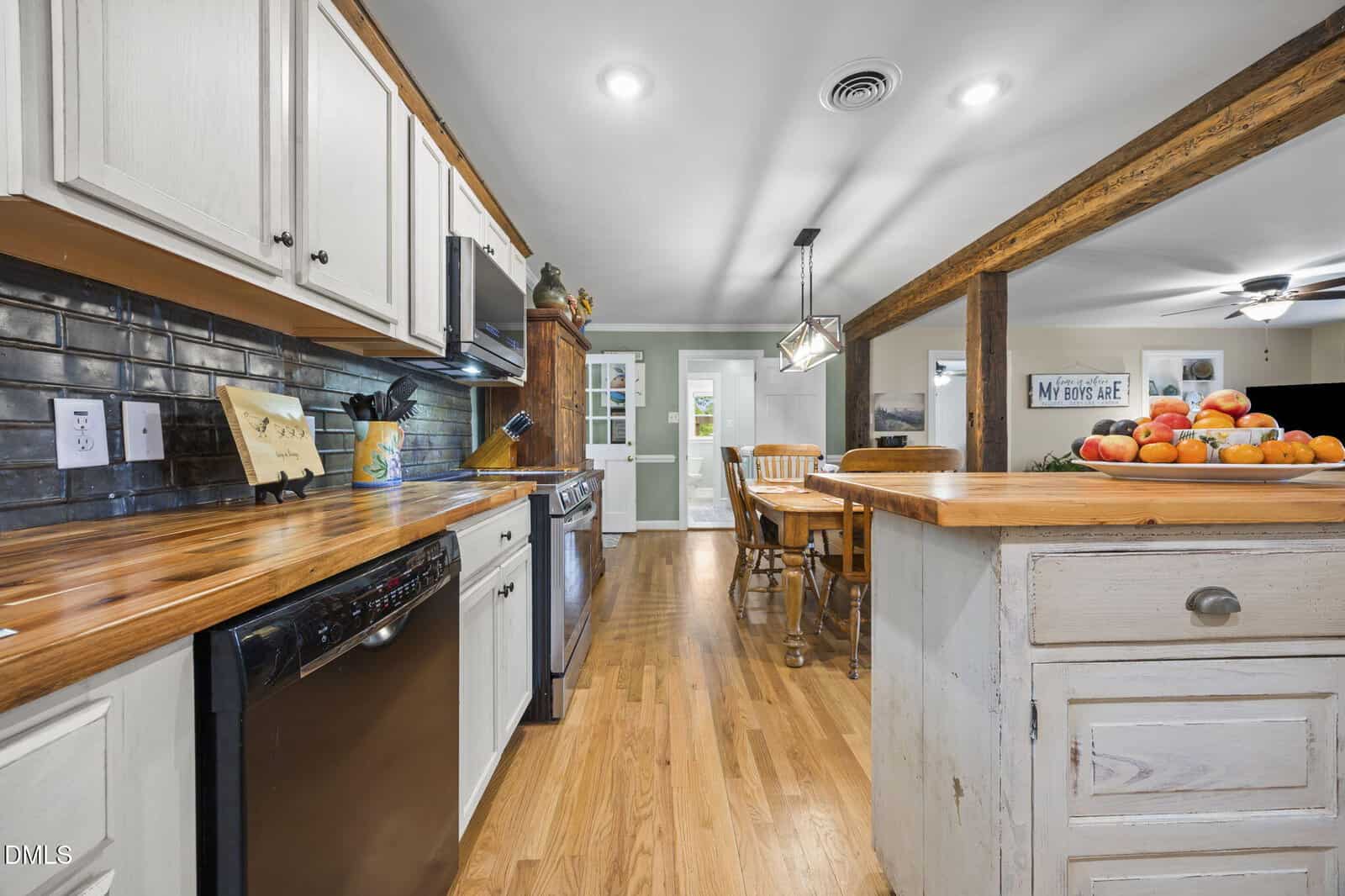 A bright kitchen with wooden floors, white cabinets, wood countertops, black appliances, a kitchen island with drawers, and a dining table with chairs. A fruit bowl sits on the island. The dining area leads to a glass door.