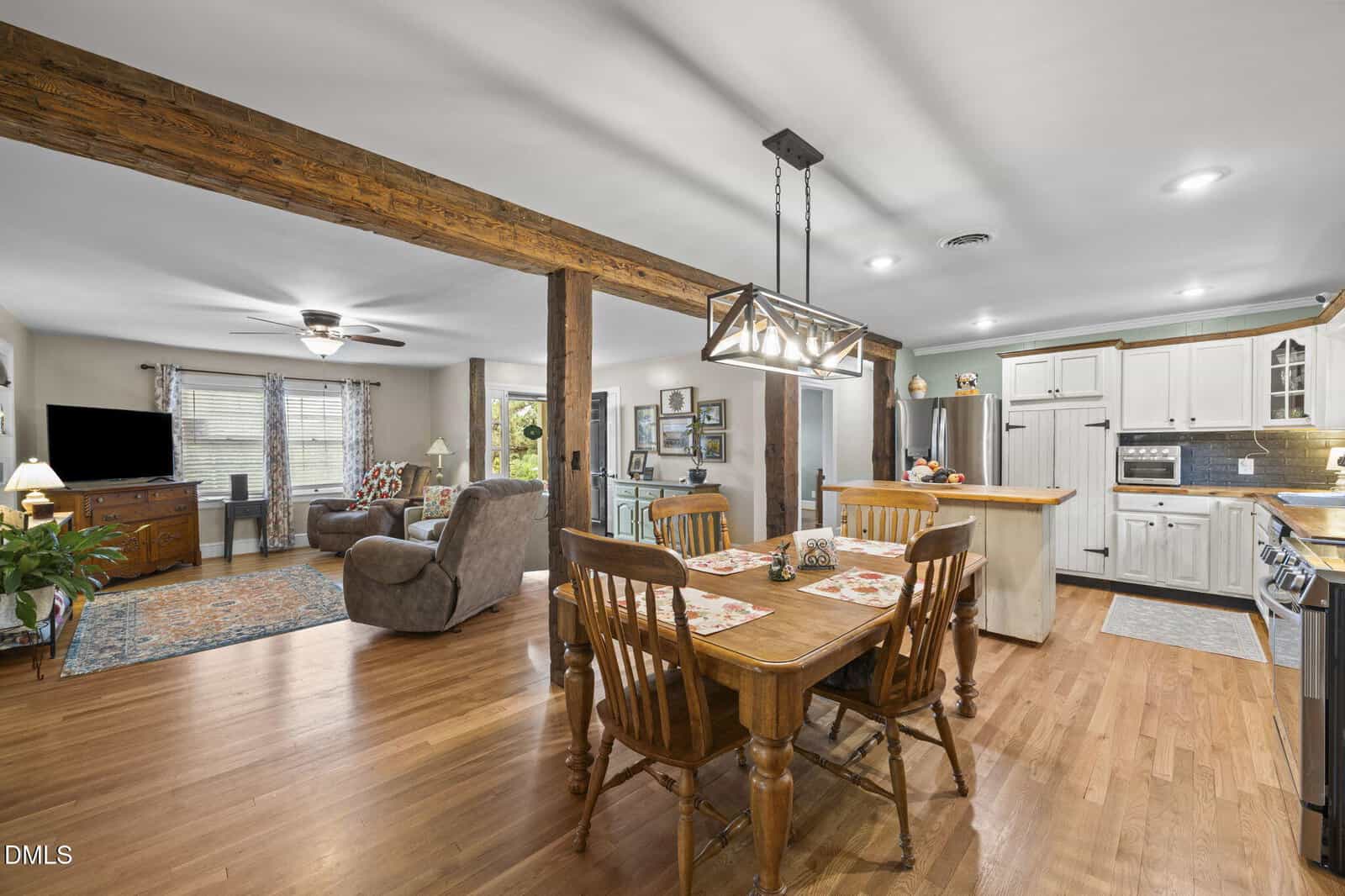 Open-concept kitchen and dining area with wood floors, wooden dining table and chairs, modern appliances, a cozy living space with a recliner and sofa, and exposed wooden beam accents. Natural light enters through large windows.