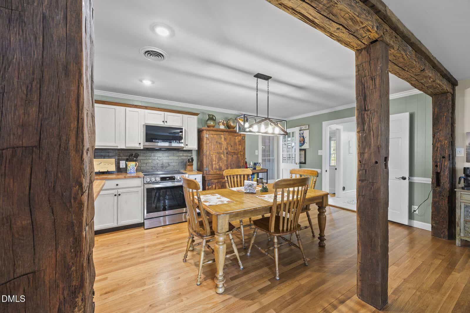 A rustic kitchen with wooden beams, light cabinets, stainless steel appliances, a wooden dining table with six chairs, and hardwood floors. Green walls and natural light give the space a cozy atmosphere.