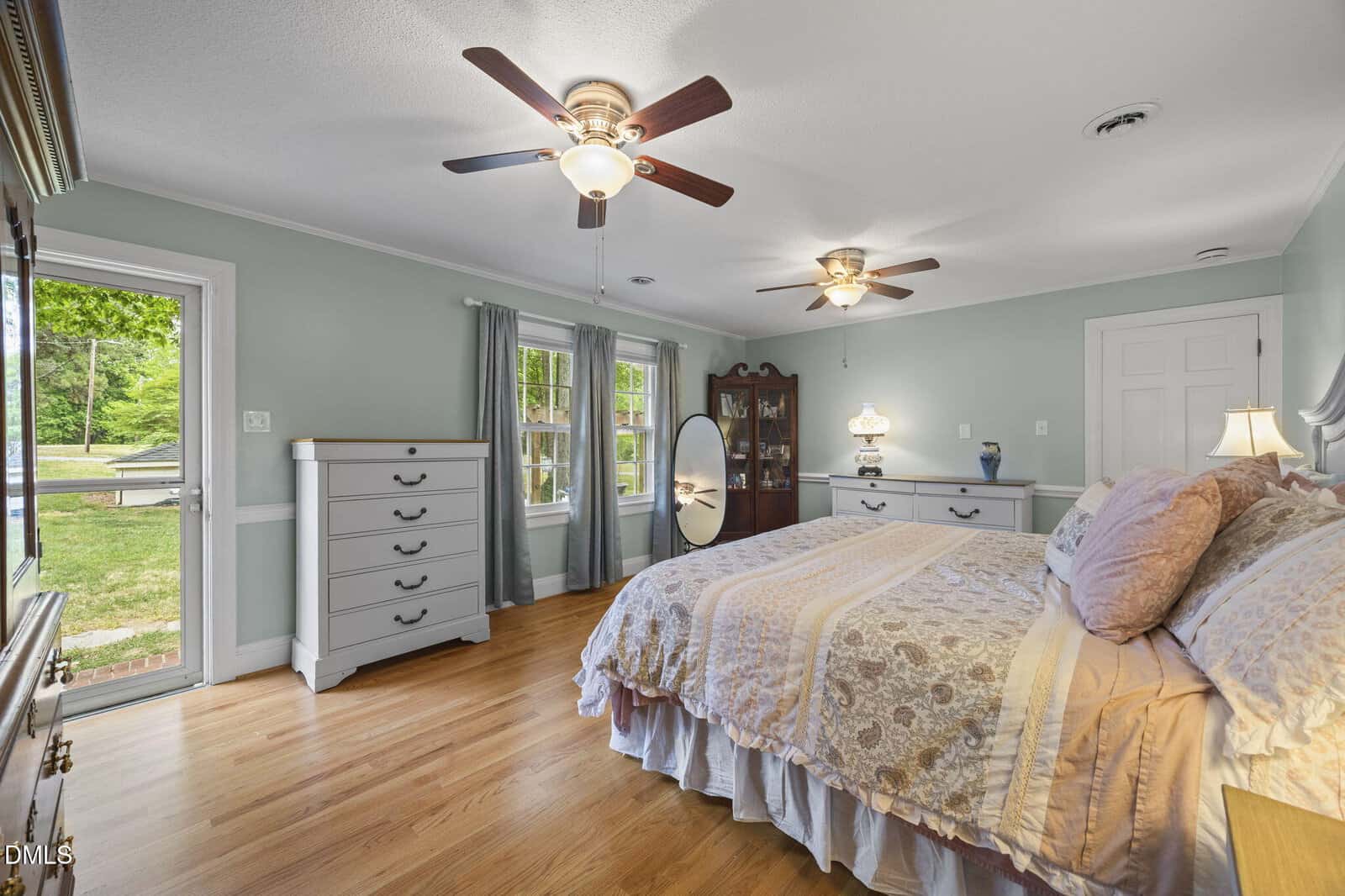 A spacious bedroom with light blue walls, hardwood floors, a large bed with patterned bedding, dressers, ceiling fans, a glass door to the outside, and several windows letting in natural light.