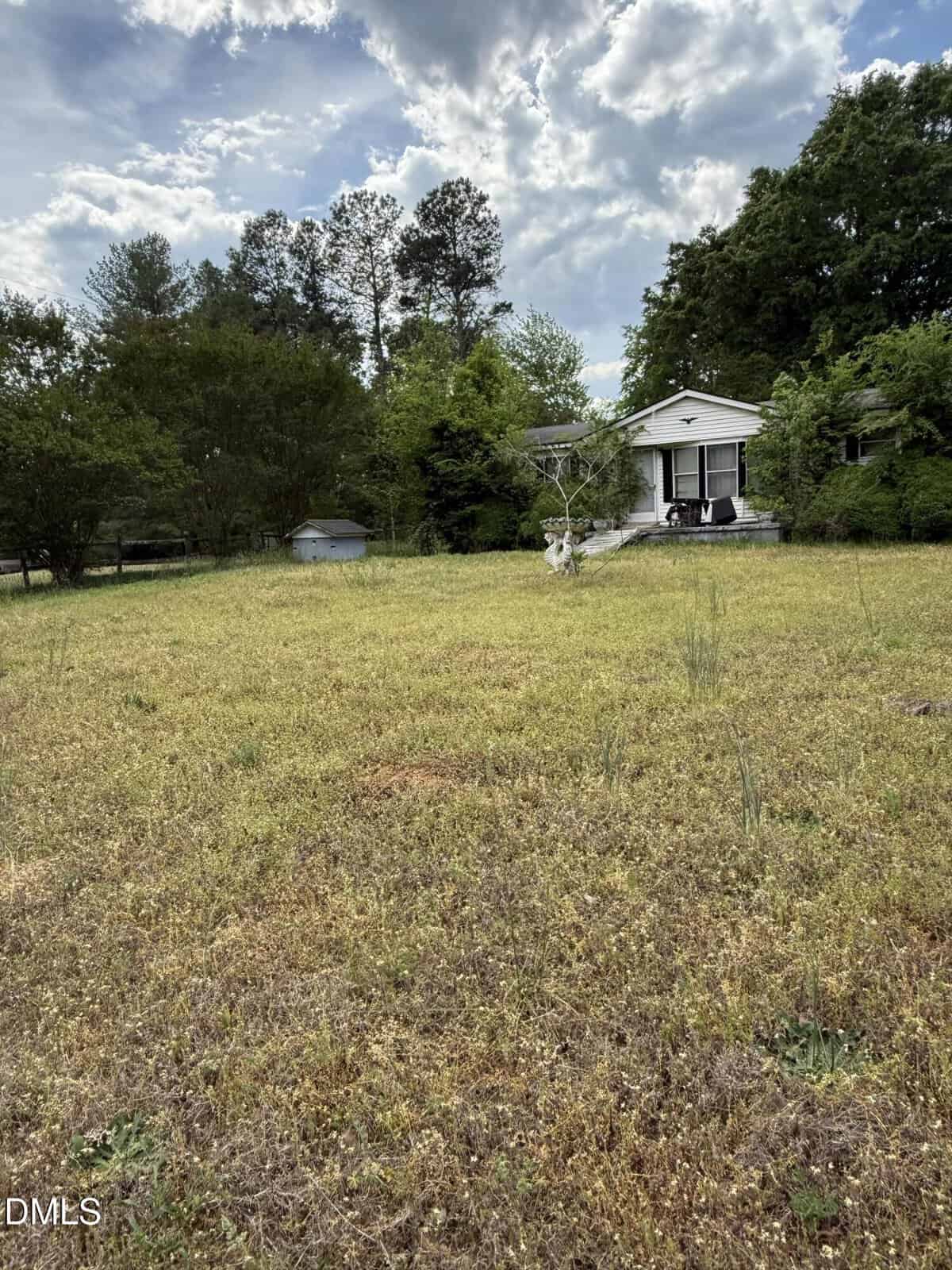 A small white house with a covered porch sits among trees and greenery, facing a large, grassy yard under a partly cloudy sky.