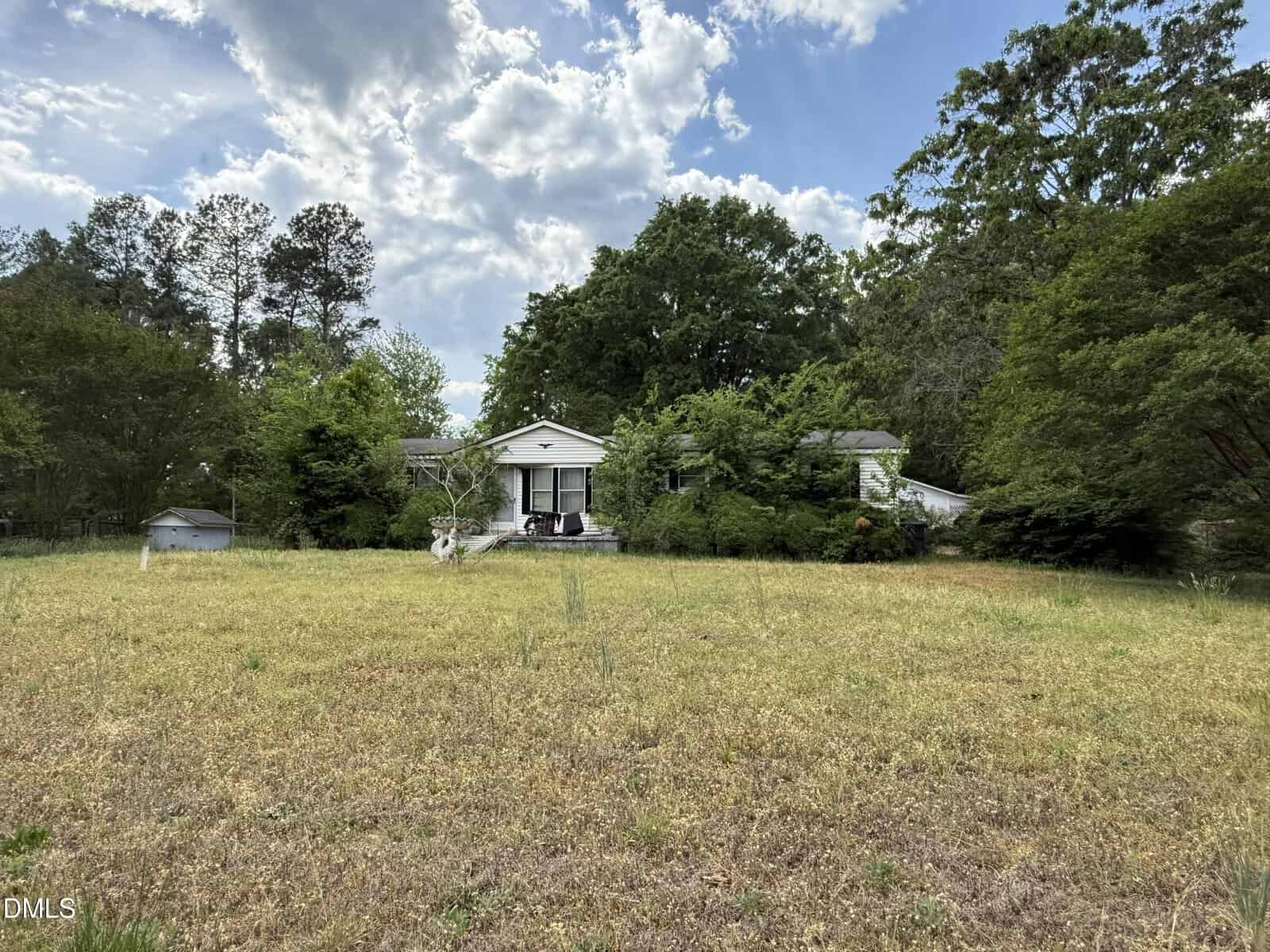 A small white house with a front porch is surrounded by trees and overgrown bushes, set back from a large grassy yard under a partly cloudy sky.