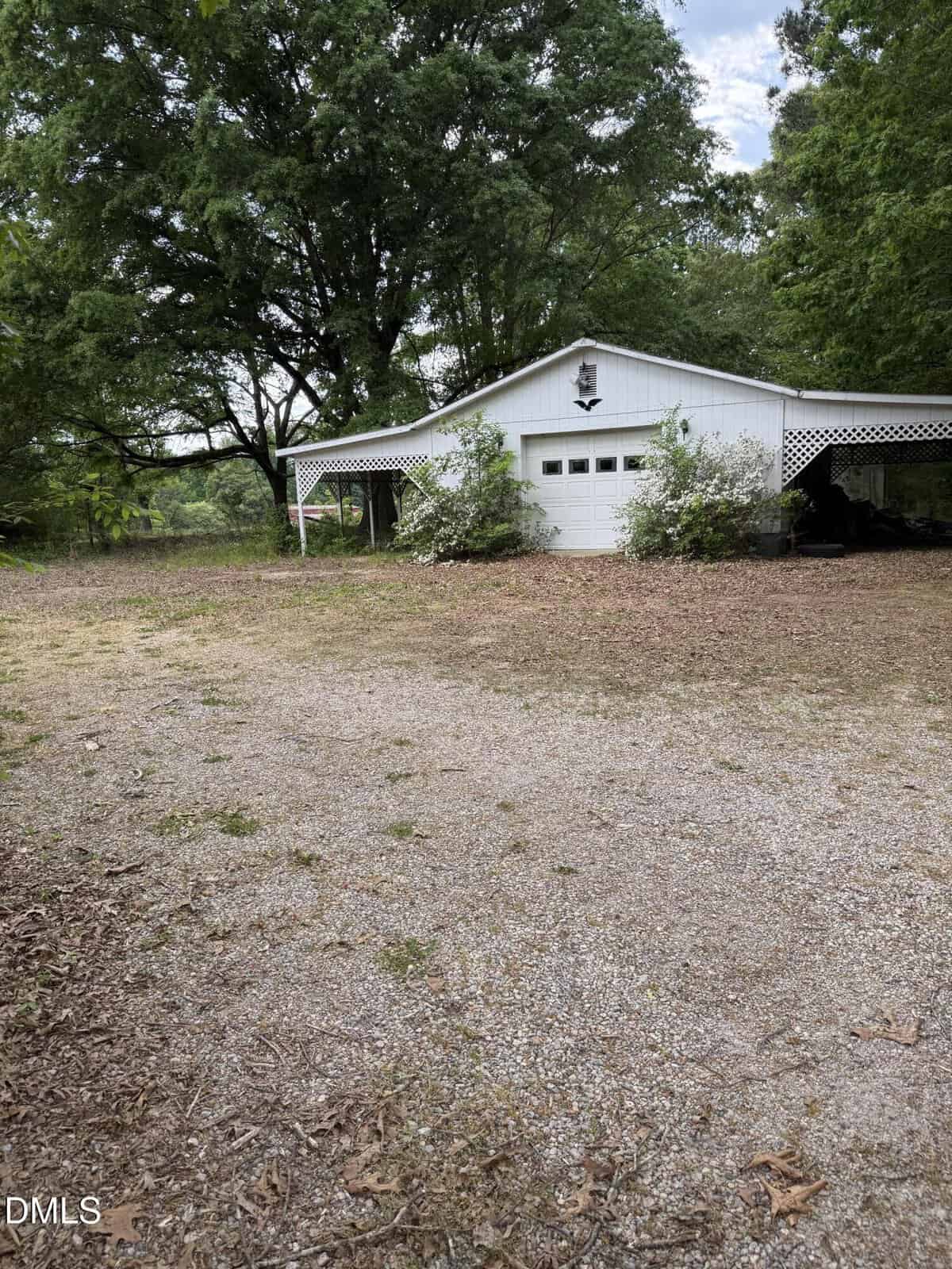 A white detached garage with a single door, carport, and overhanging trees. The driveway is gravel with scattered leaves, and there is greenery around the building.