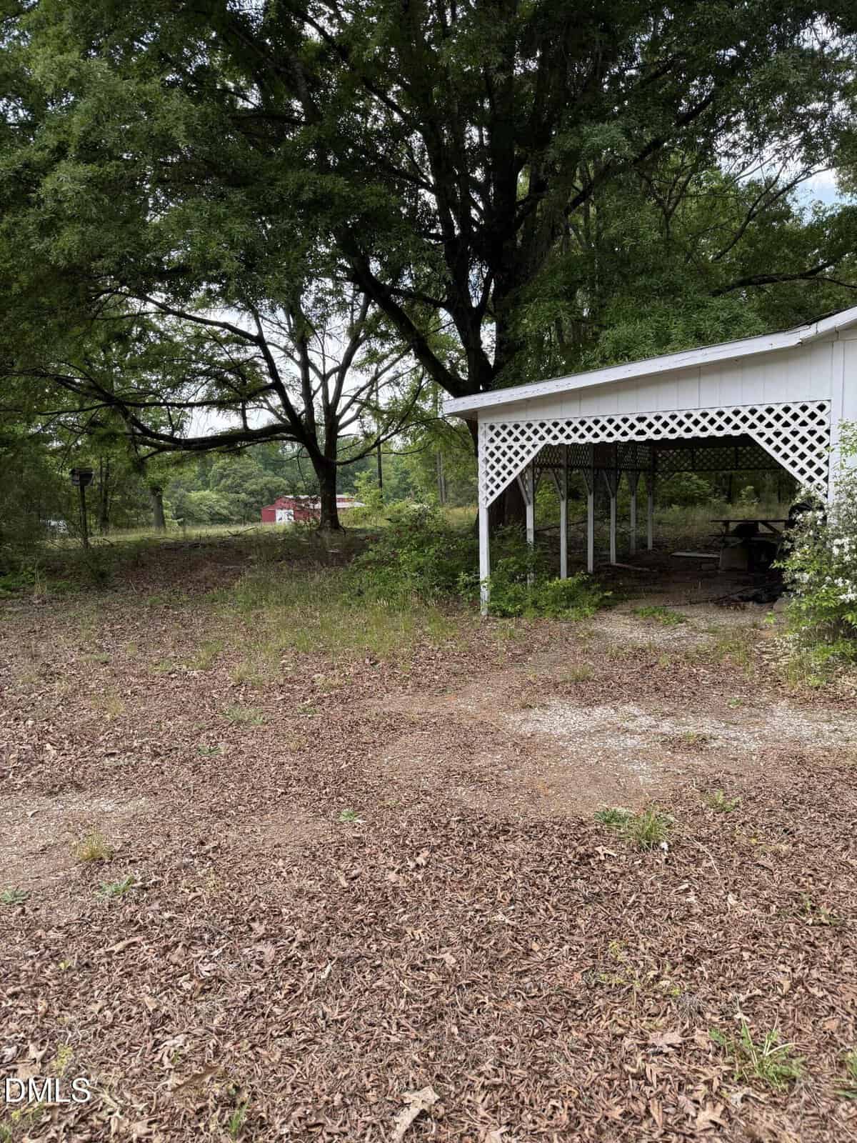 A white carport with lattice trim stands beside a dirt and leaf-covered yard, surrounded by tall trees and greenery. A red building is visible in the background.