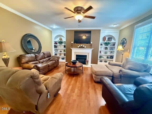 A cozy living room with brown leather sofas and armchairs, a fireplace with a TV above it, built-in white shelves with decor, hardwood floors, and a ceiling fan. Large window on the right lets in natural light.