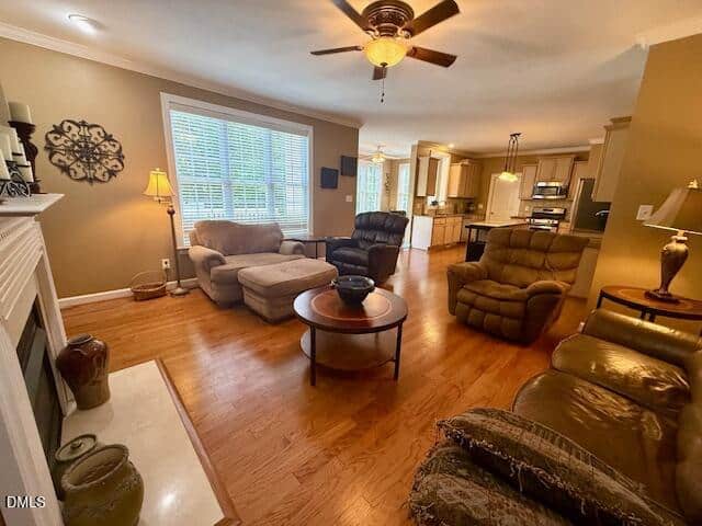 Spacious living room with brown leather sofas, armchairs, coffee tables, hardwood floors, a ceiling fan, large window, fireplace, and an open view into a modern kitchen.