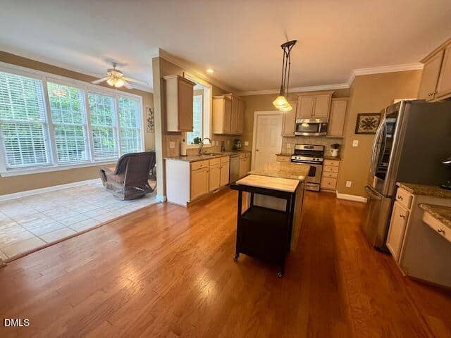 Spacious kitchen with light wood cabinets, stainless steel appliances, center island, and hardwood floors. Attached sunroom has large windows, tile flooring, a ceiling fan, and a single armchair. Warm lighting throughout.