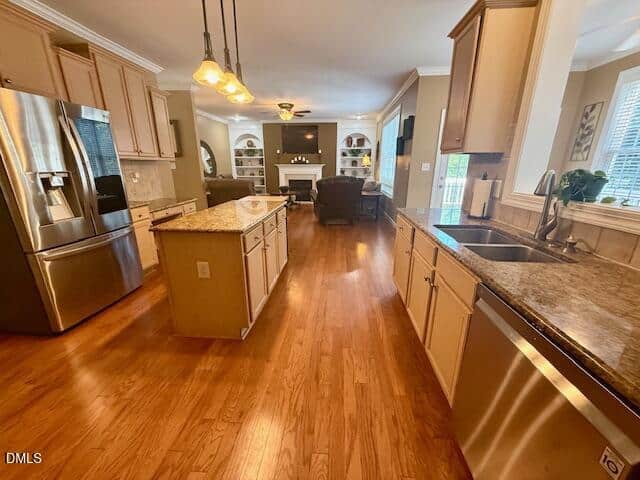 Modern kitchen with light wood cabinets, granite countertops, stainless steel appliances, island, and open view into a cozy living room with hardwood floors, fireplace, armchairs, and built-in shelves by large windows.