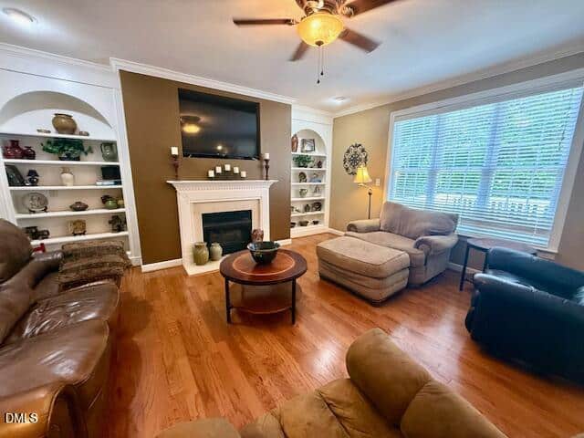Living room with brown leather sofas, a fireplace under a wall-mounted TV, built-in shelves with decor, wood floors, large window with blinds, and a ceiling fan with lights.