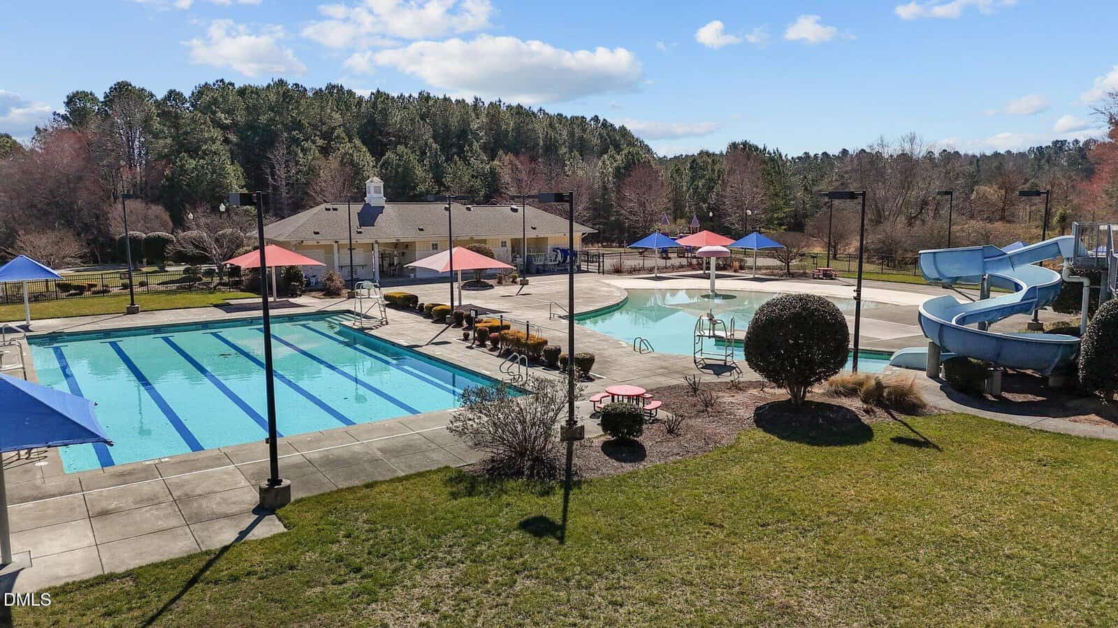 Outdoor swimming pool complex with a lap pool, a curvy water slide, and a separate pool area; several red and blue shade umbrellas and lounge chairs; surrounded by grass, trees, and a clubhouse under a partly cloudy sky.