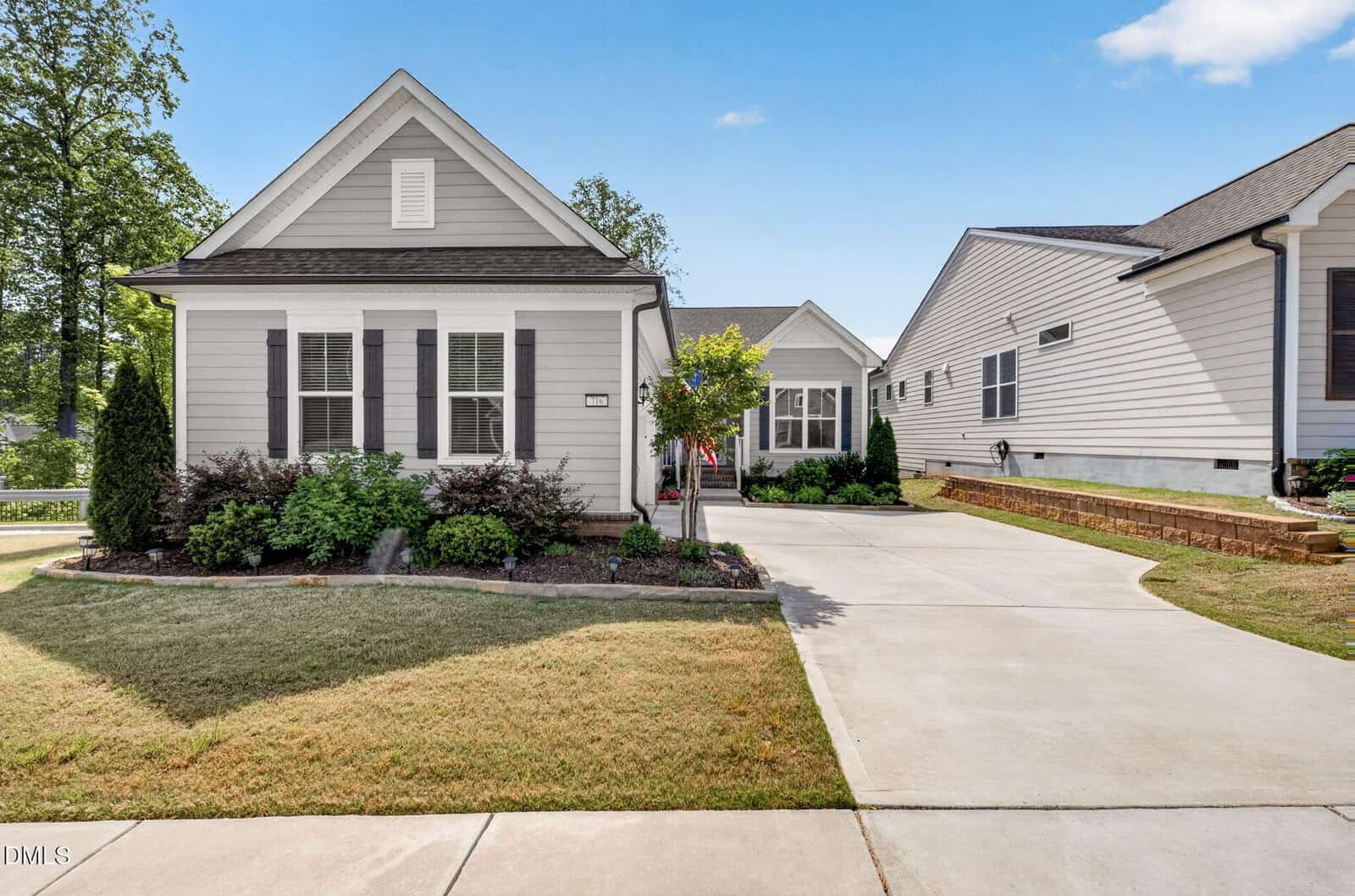 A single-story house with gray siding, white trim, and a gable roof, featuring a neatly landscaped front yard, shrubs, and a concrete driveway leading to a garage on a sunny day.