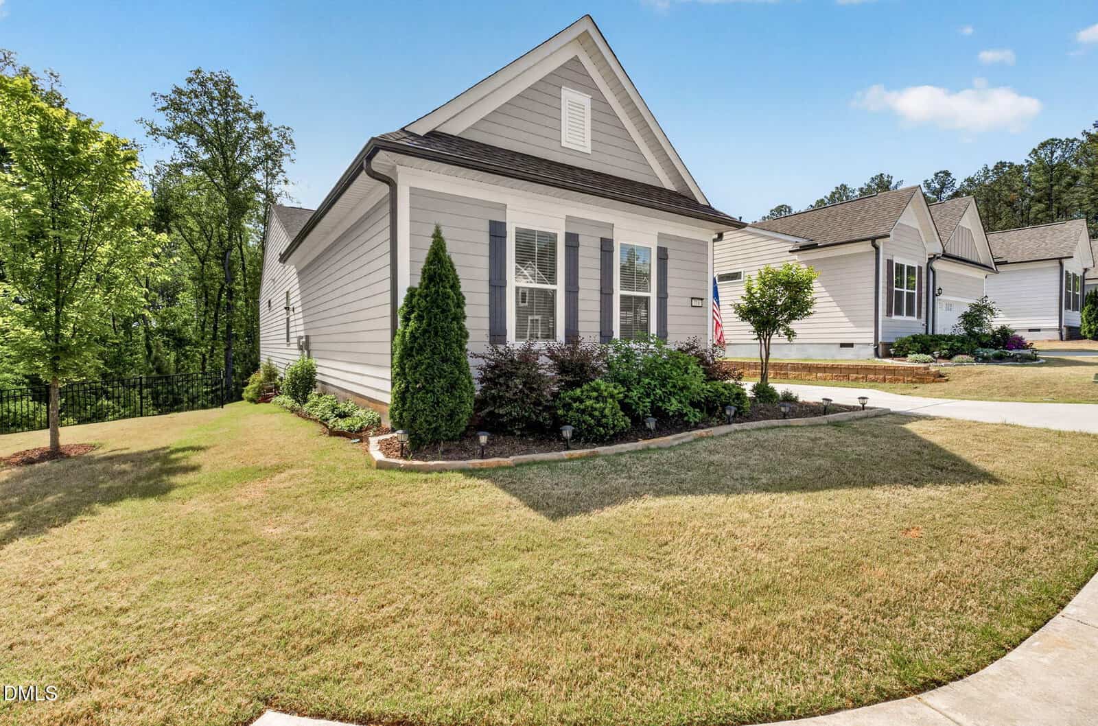 Single-story light gray house with white trim, small covered entry, and black shutters. Neatly landscaped yard with shrubs and mulch, set on a corner lot with a curved driveway and a well-kept lawn. Trees in the background.
