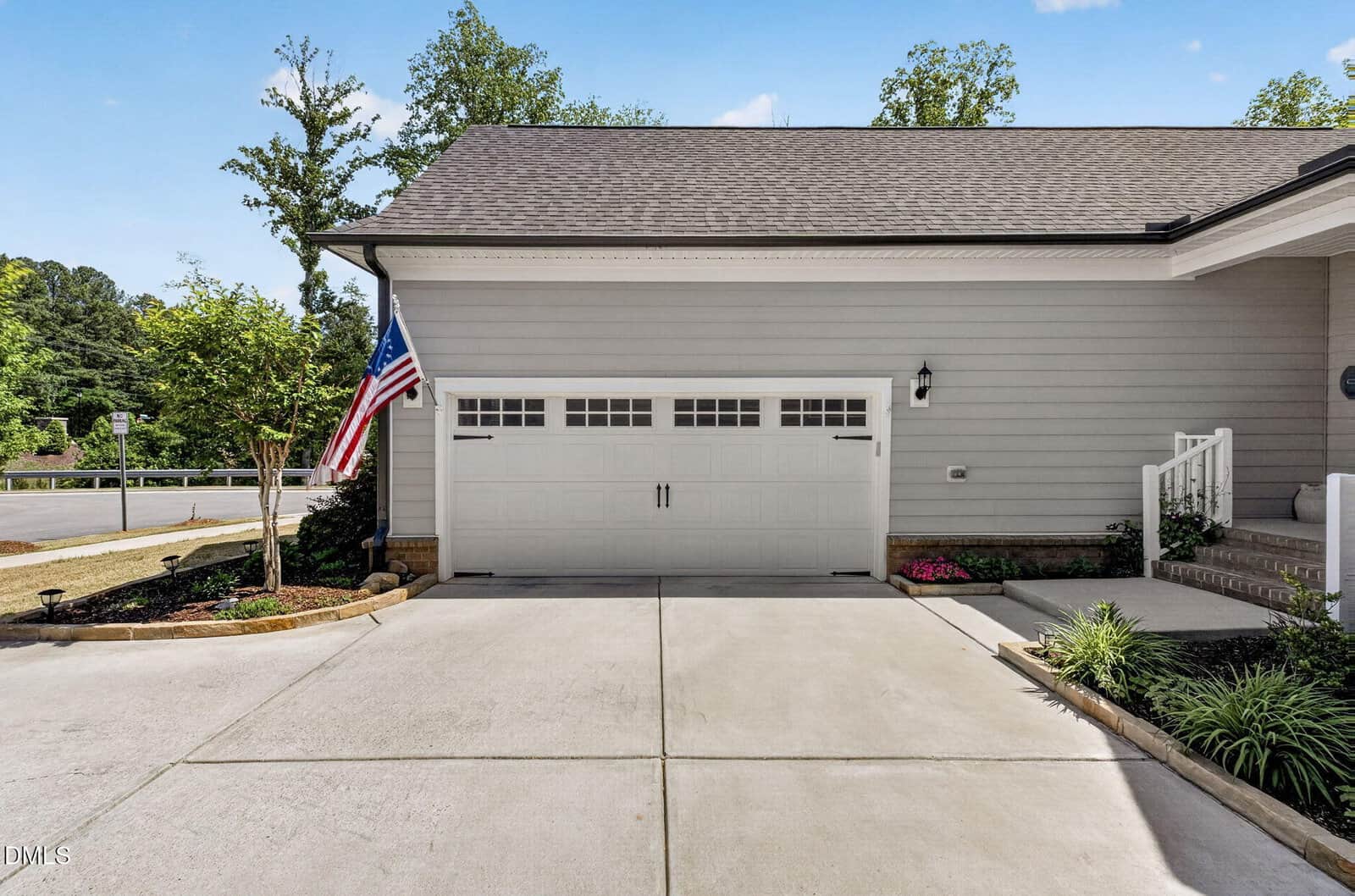 A modern suburban house with light gray siding, a white double garage door with windows, an American flag, landscaped plants, and a concrete driveway on a sunny day.