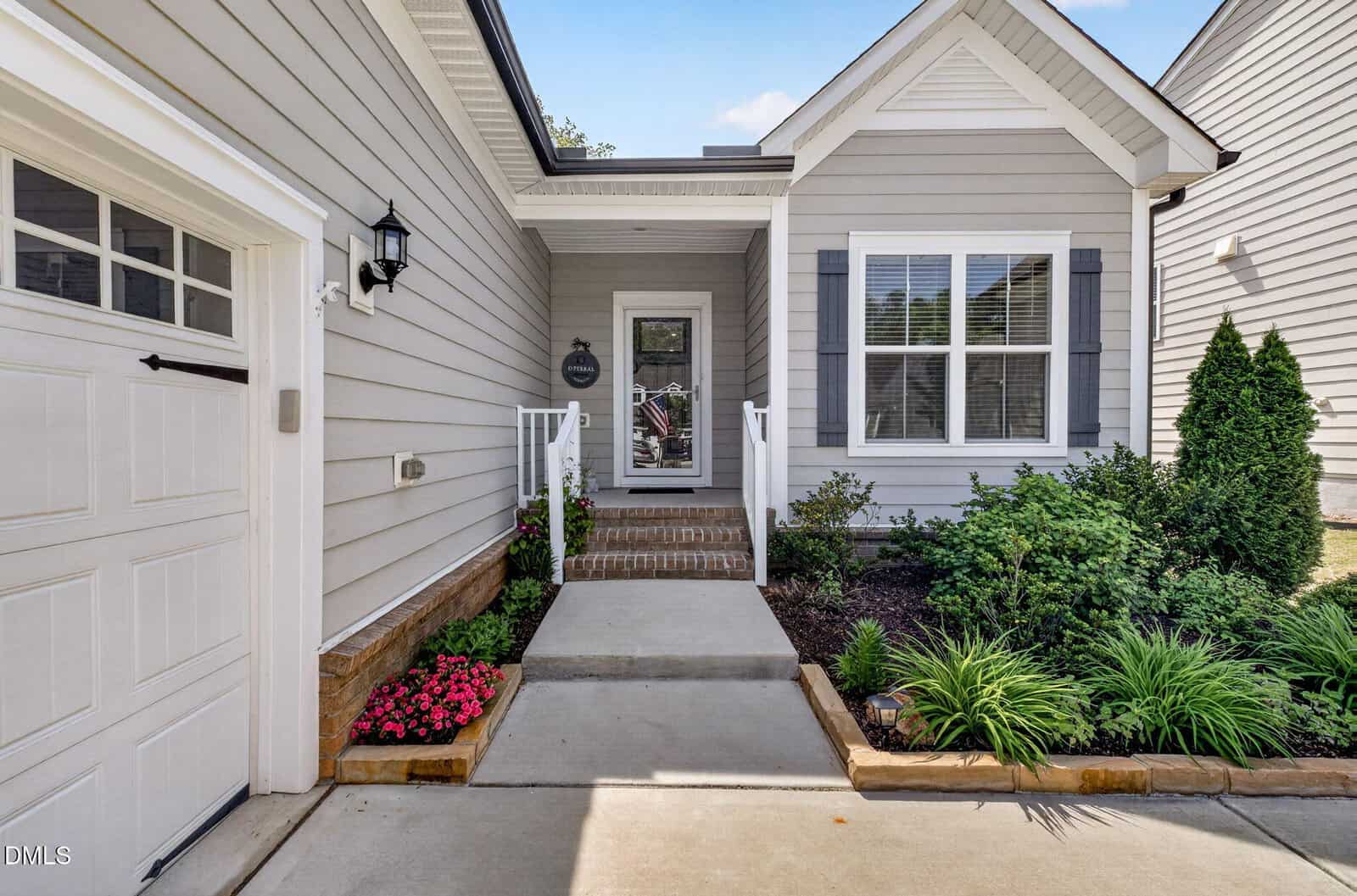 A concrete walkway leads to the front door of a gray house with white trim, large windows with shutters, a small porch, and neatly landscaped garden beds with green plants and pink flowers.