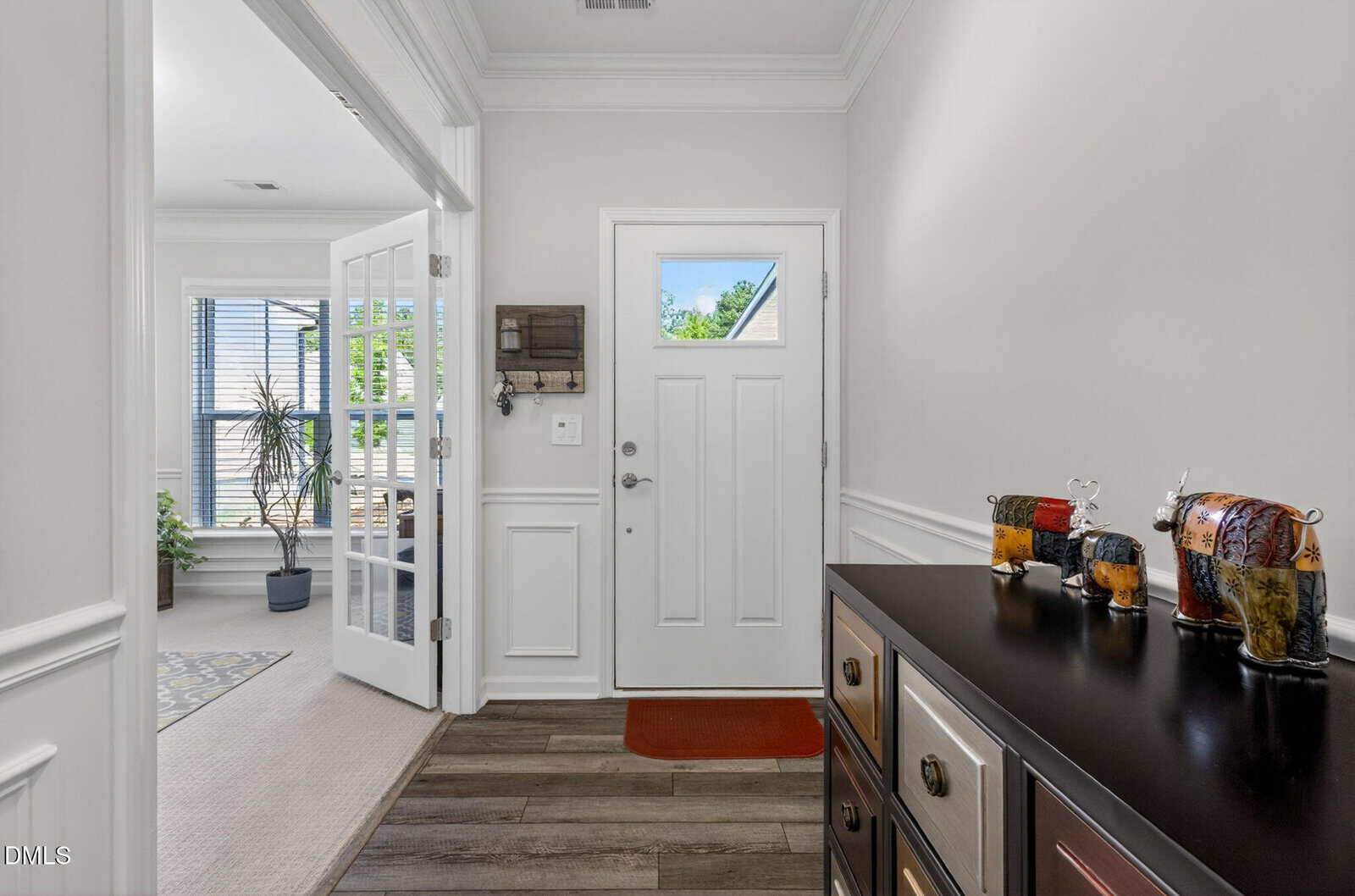 A bright entryway with a white front door, wood flooring, a black sideboard with colorful animal sculptures, and a room with large windows and a tall plant visible through French doors.