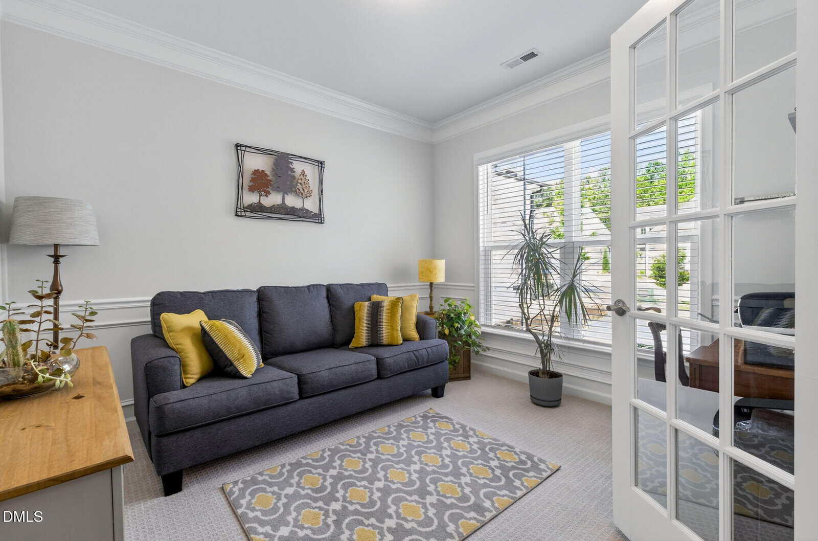 Bright home office with gray sofa, yellow accents, large window with blinds, indoor plants, wooden side table, modern floor lamp, wall art, and glass French doors. Natural light fills the room.