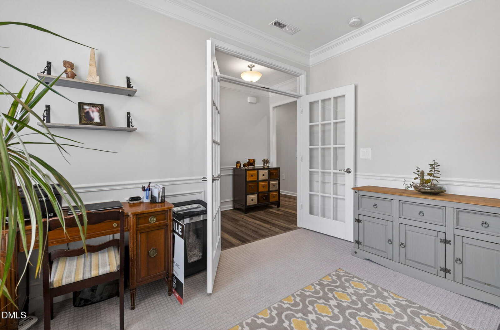 Home office with a wooden desk and chair, shelves with decor, a gray sideboard, and a yellow-patterned rug. French doors open to a hallway with wooden flooring and a cabinet with drawers.