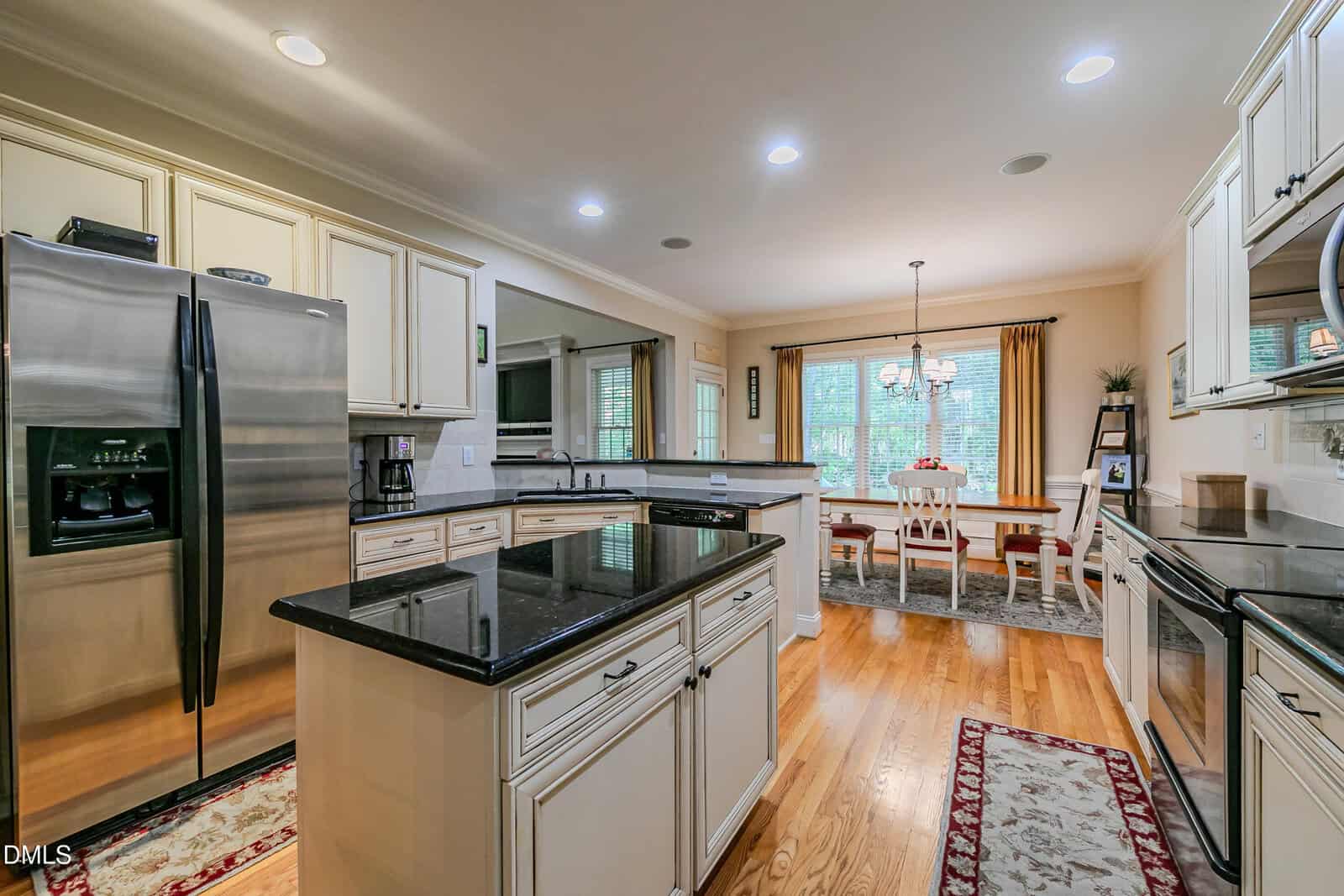 Modern kitchen with cream cabinets, black countertops, stainless steel appliances, and wooden floors. An island is centered, and a dining area with a window and chandelier is visible in the background.