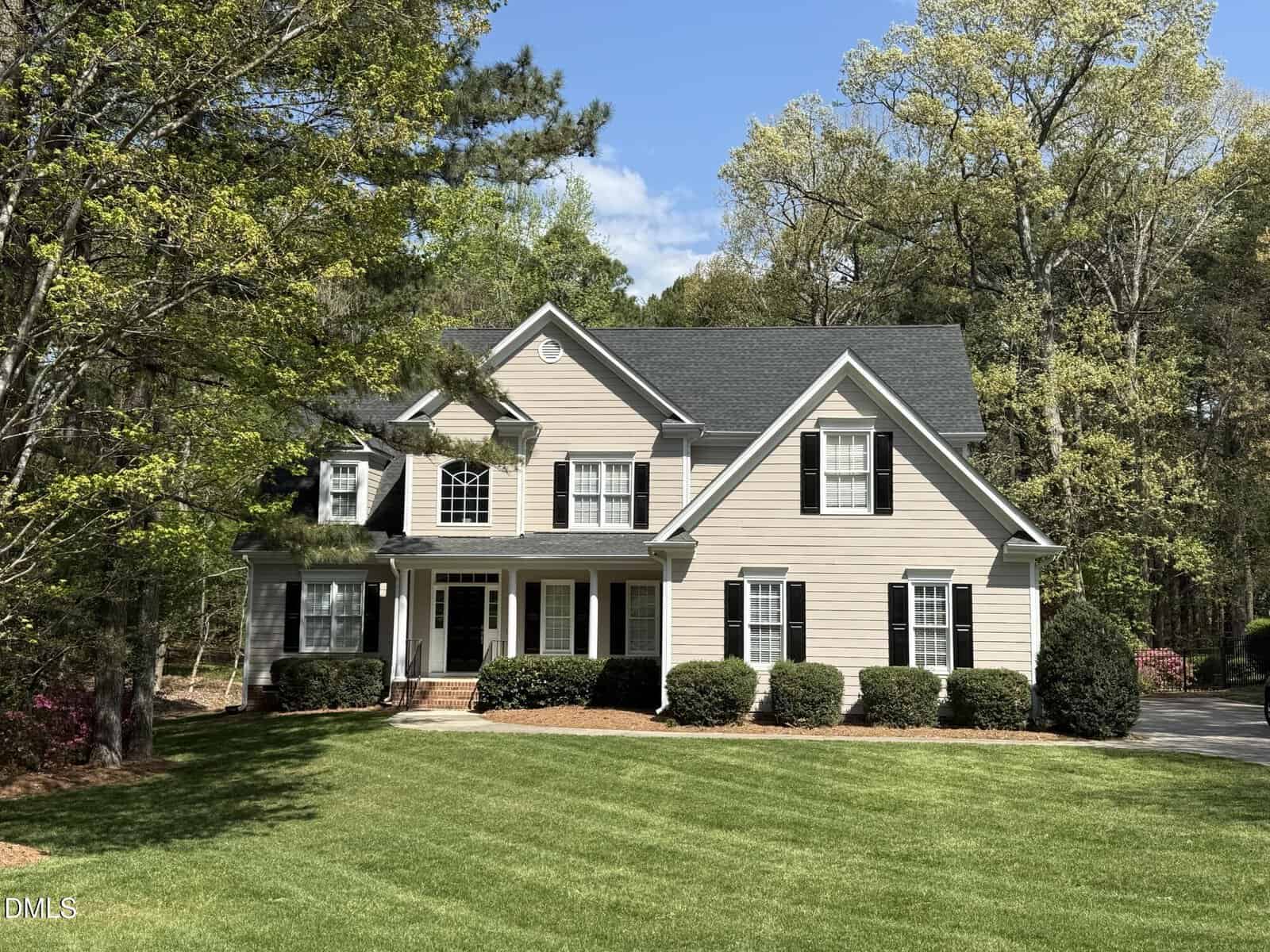 The two-story beige house at 140 Traylee Drive, Wake Forest, features white trim, black shutters, a dark roof, and is surrounded by green lawns, bushes, and tall trees under a partly cloudy sky.