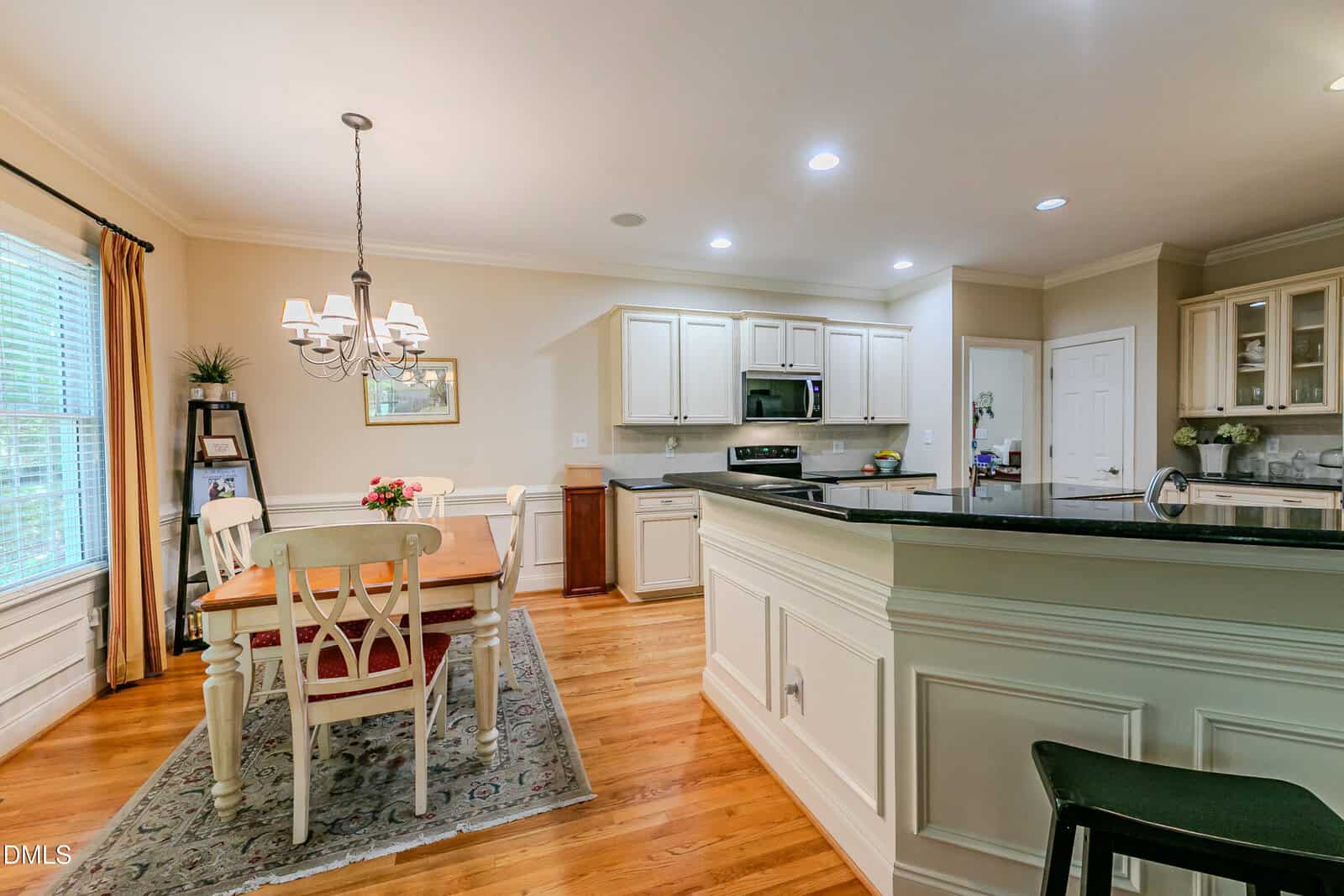 Bright kitchen and dining area with hardwood floors, white cabinets, stainless steel appliances, a chandelier over a wooden table, and sunlight streaming in through large windows.