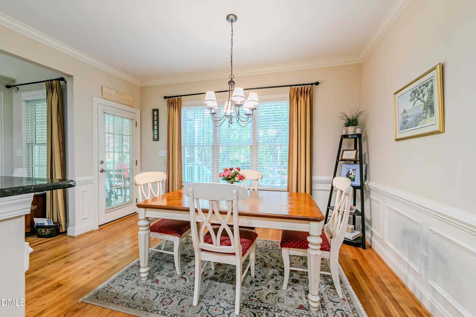 A bright dining room with a wooden table, five chairs, a rug, chandelier, large window with blinds, and beige walls adorned with art and a shelving unit in the corner.