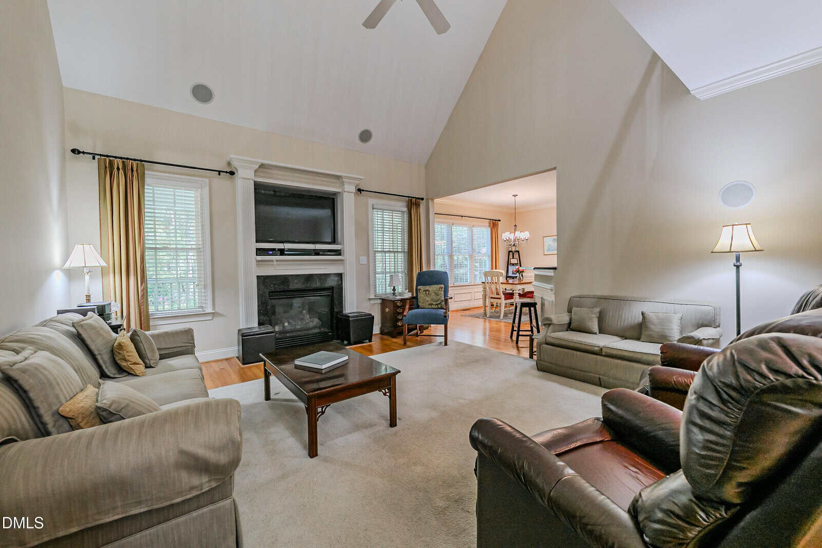 Spacious living room with vaulted ceiling, neutral walls, sofas, armchairs, coffee table, lamps, a fireplace with a TV above, and large windows; open view into dining area and kitchen.