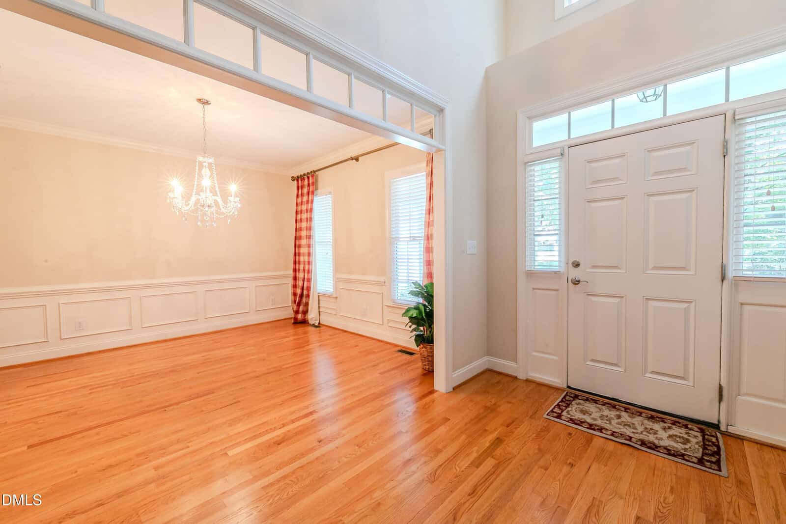 Spacious entryway with light wood floors, a white front door with sidelights, and an adjacent dining room featuring large windows, a chandelier, and red curtains. The space is bright and open with white trim.