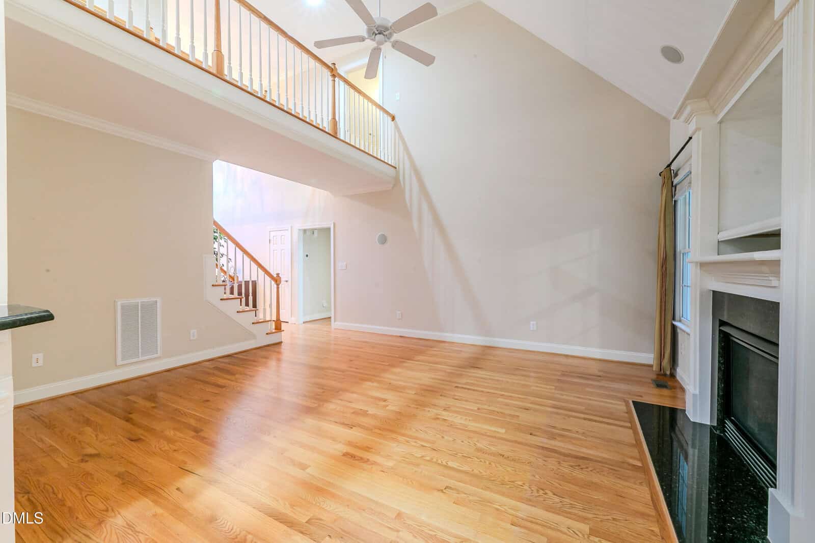 Spacious living room with high vaulted ceiling, ceiling fan, light wood floors, a staircase with wooden railings, large windows, and a white fireplace with a black hearth on the right wall.