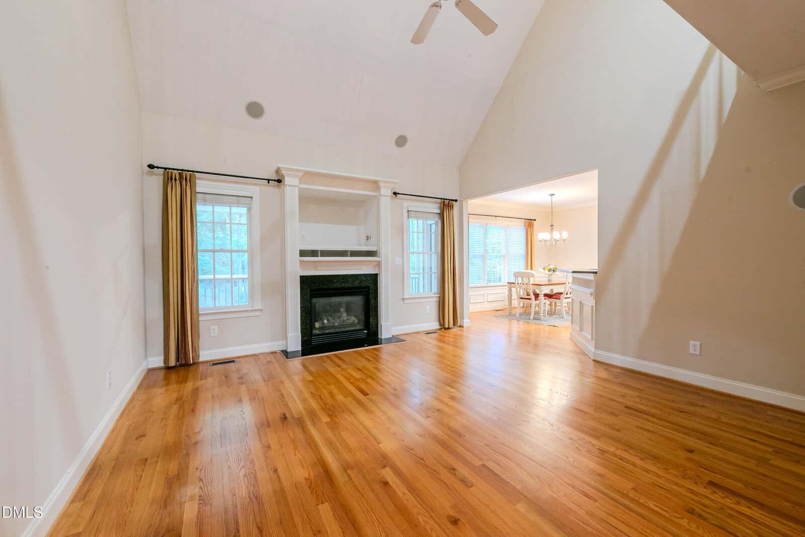 Spacious living room with hardwood floors, a black fireplace with white mantel, built-in shelves, two large windows, and an open view into a bright dining area with a chandelier.
