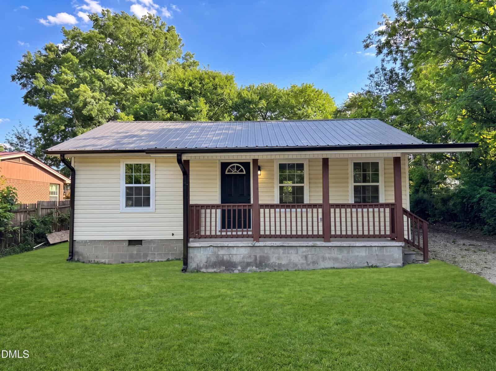 Single-story house with cream siding, a dark metal roof, and a covered front porch with wooden railings. The home sits on a fresh green lawn, surrounded by trees under a clear blue sky.