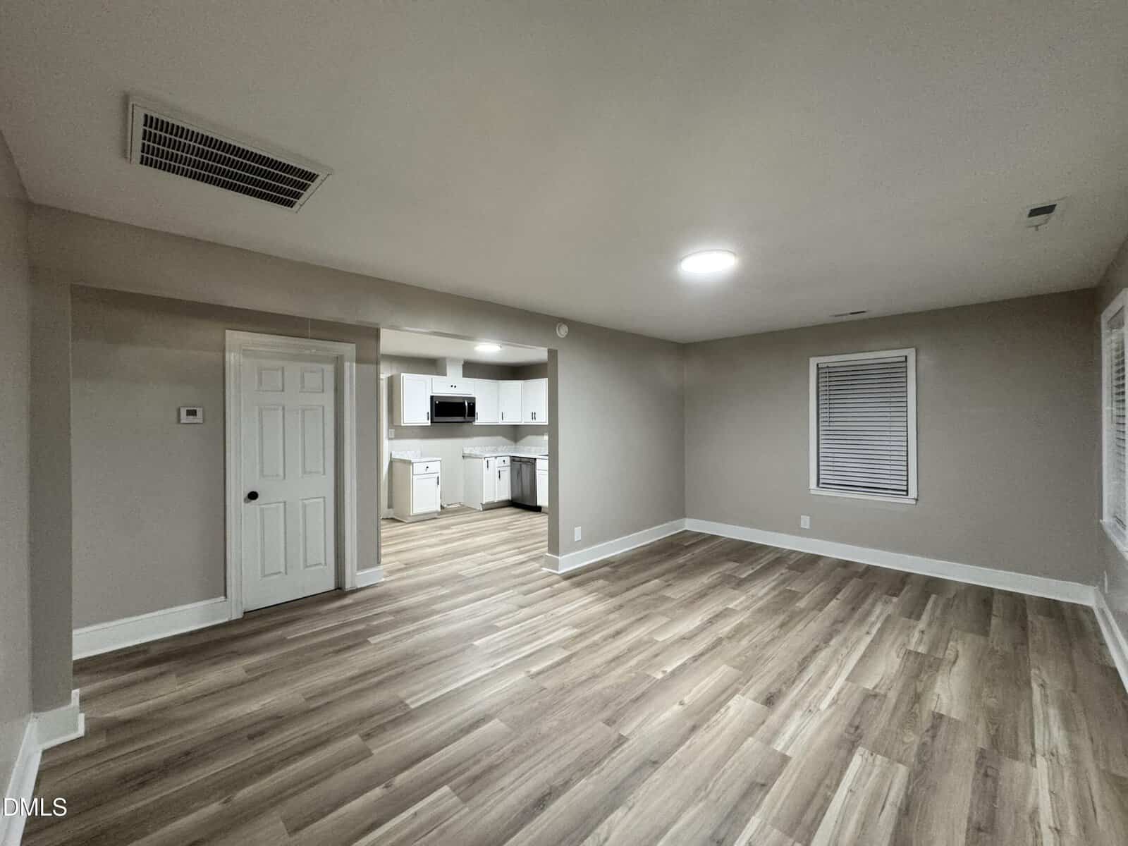 Spacious, empty room with light wood floors and beige walls, leading to a white kitchen with cabinets and appliances. A closed door and two windows are visible; ceiling lights brighten the space.