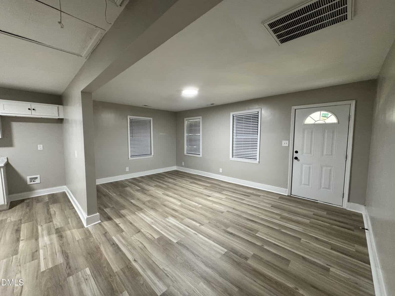 A spacious, empty room with light wood flooring, beige walls, three windows with white blinds, a white entry door, and a ceiling vent. The room appears clean and well-lit.