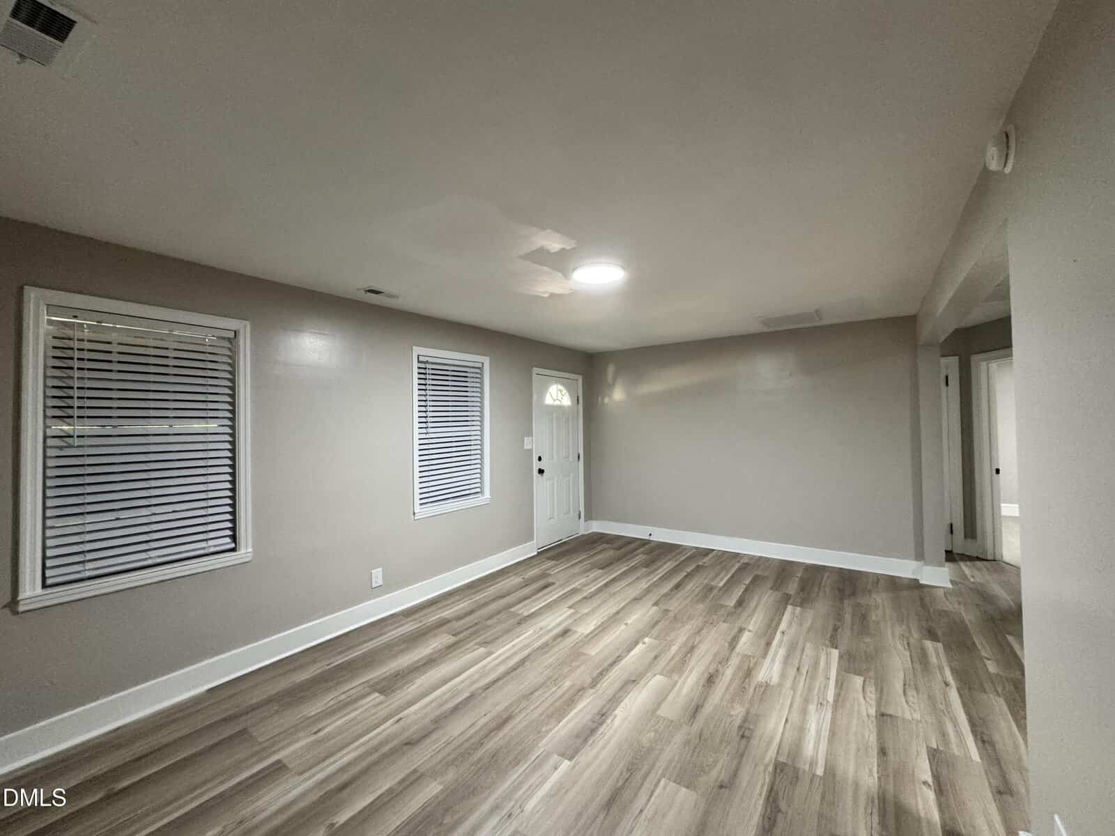 A bright, empty room with light wood flooring, beige walls, two windows with blinds, and a white front door. There is a circular ceiling light and a patch on the ceiling.