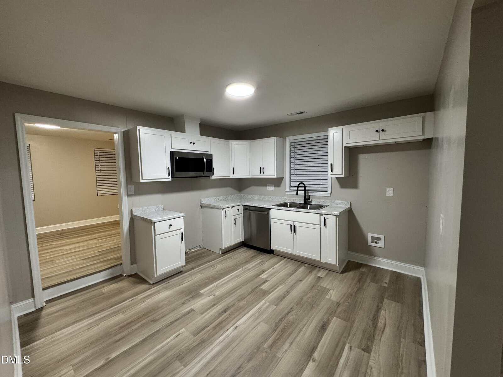 Modern kitchen with white cabinets, marble countertops, wood-style flooring, a sink under a window, built-in microwave, dishwasher, and neutral-colored walls. An open doorway leads to another room.