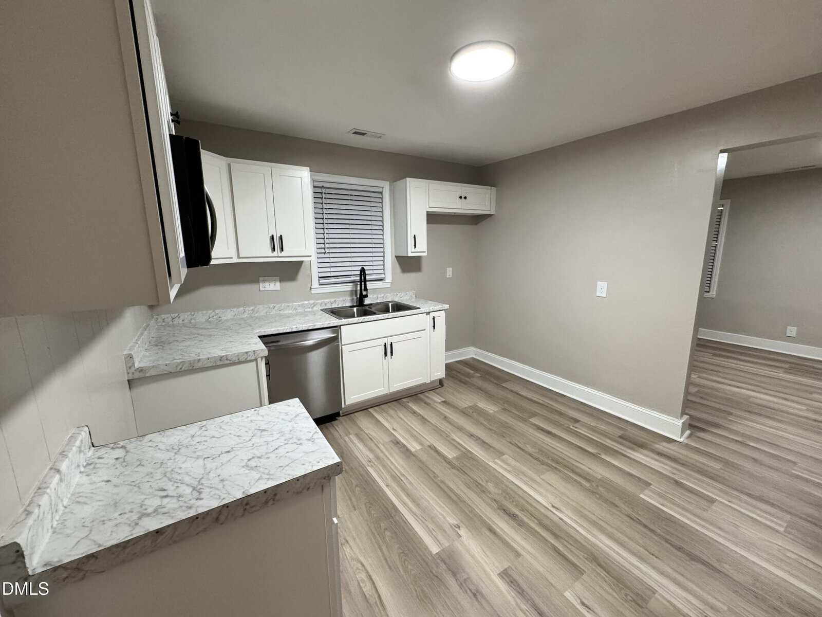Modern kitchen with light wood flooring, white cabinets, marble countertops, stainless steel dishwasher, sink under two windows, and beige walls. Room has recessed lighting and an open doorway to another space.