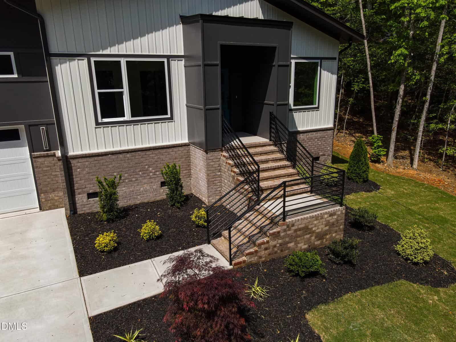 A modern house entrance with brick stairs, black metal railings, and landscaped garden beds featuring shrubs and mulch, adjacent to a concrete driveway and green lawn.