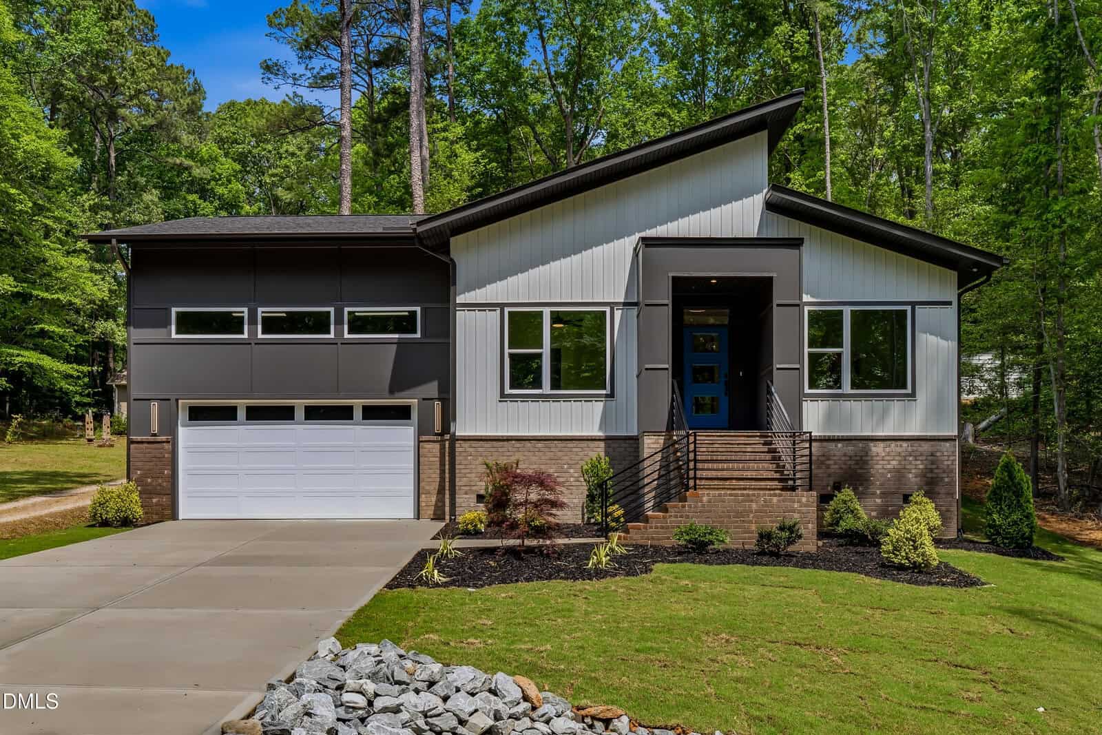 Modern two-story house with a sloped roof, large windows, gray and white exterior, brick accents, and a two-car garage, surrounded by green lawn, landscaped plants, and trees in the background.