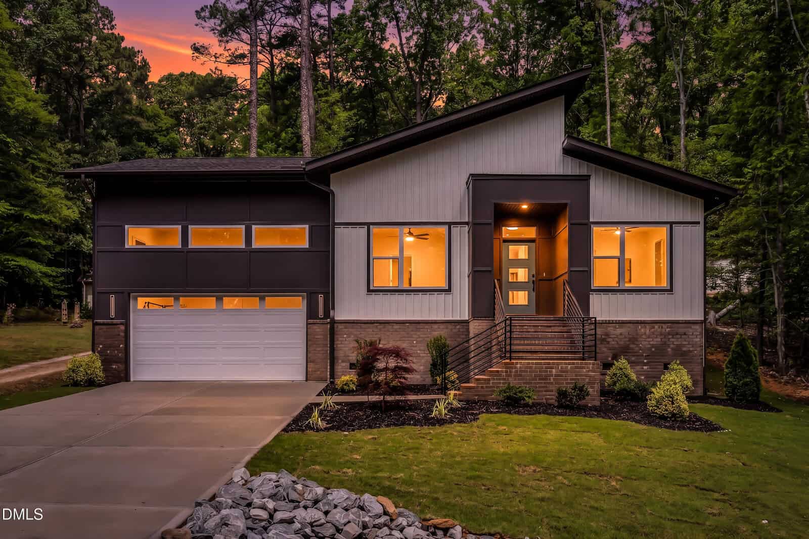 Modern house at sunset with large windows warmly lit from inside, a two-car garage, front steps leading to a glass door, landscaped yard, and surrounded by tall trees and greenery.
