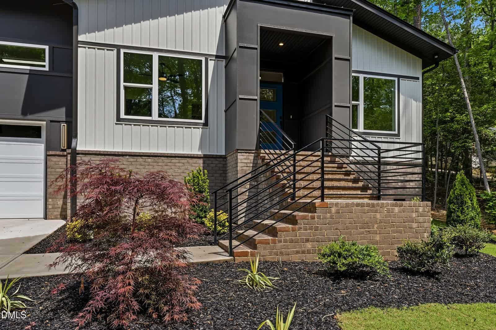 Modern house exterior with brick steps, black metal railing, and landscaped front yard featuring mulch, small bushes, and a red Japanese maple tree. Large windows and a covered entryway are visible.