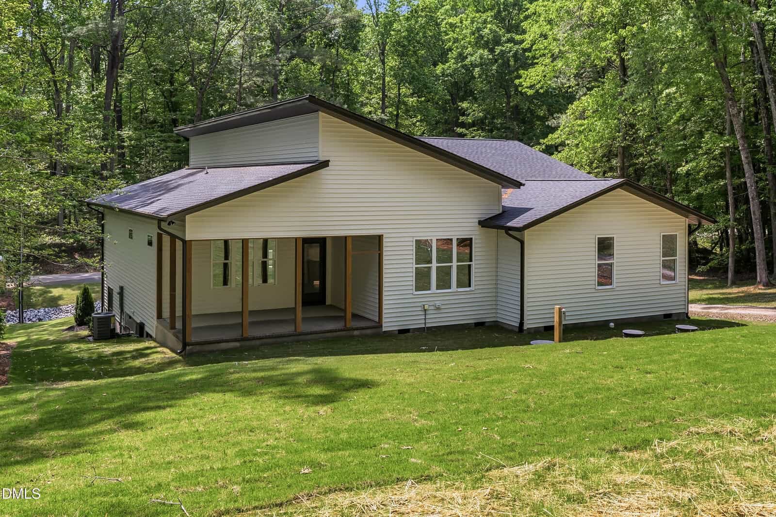 Modern single-story house with light-colored siding, large windows, and a covered porch, surrounded by green grass and trees in a wooded area.