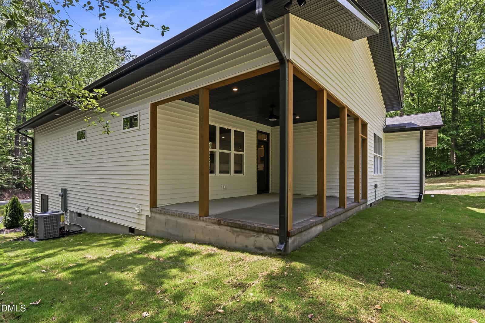 A modern house exterior with light siding, wood posts, covered patio, and windows. The yard is grassy and surrounded by trees. The scene is bright and sunny.