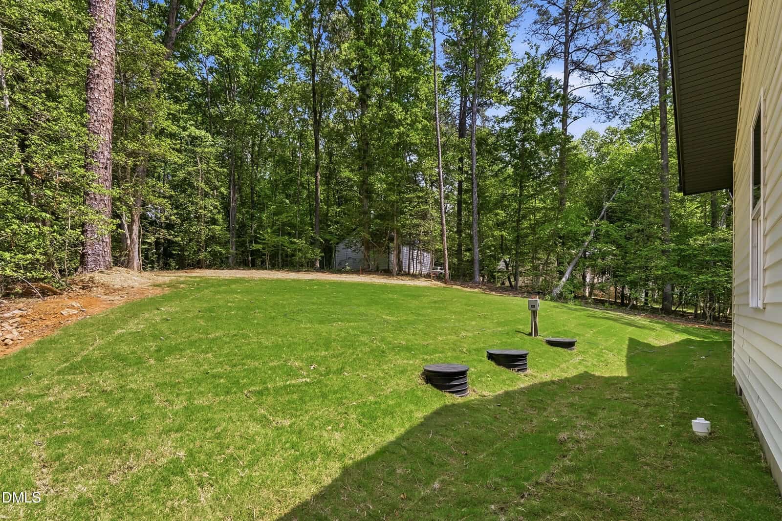 A grassy backyard slopes toward a wooded area. Three black drainage pipes are partially buried in the lawn near the yellow-sided house, which is partially visible at the right edge. Dense trees fill the background.