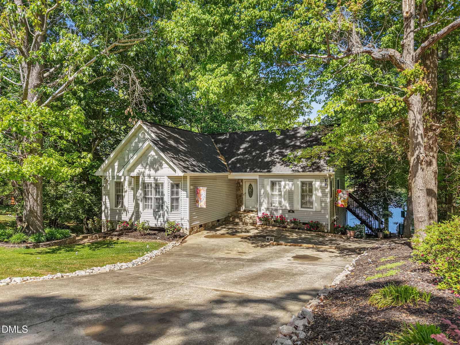 A light gray single-story house with white trim, surrounded by lush green trees. A curved driveway leads to the entrance, and there's a glimpse of water visible through the trees in the background.