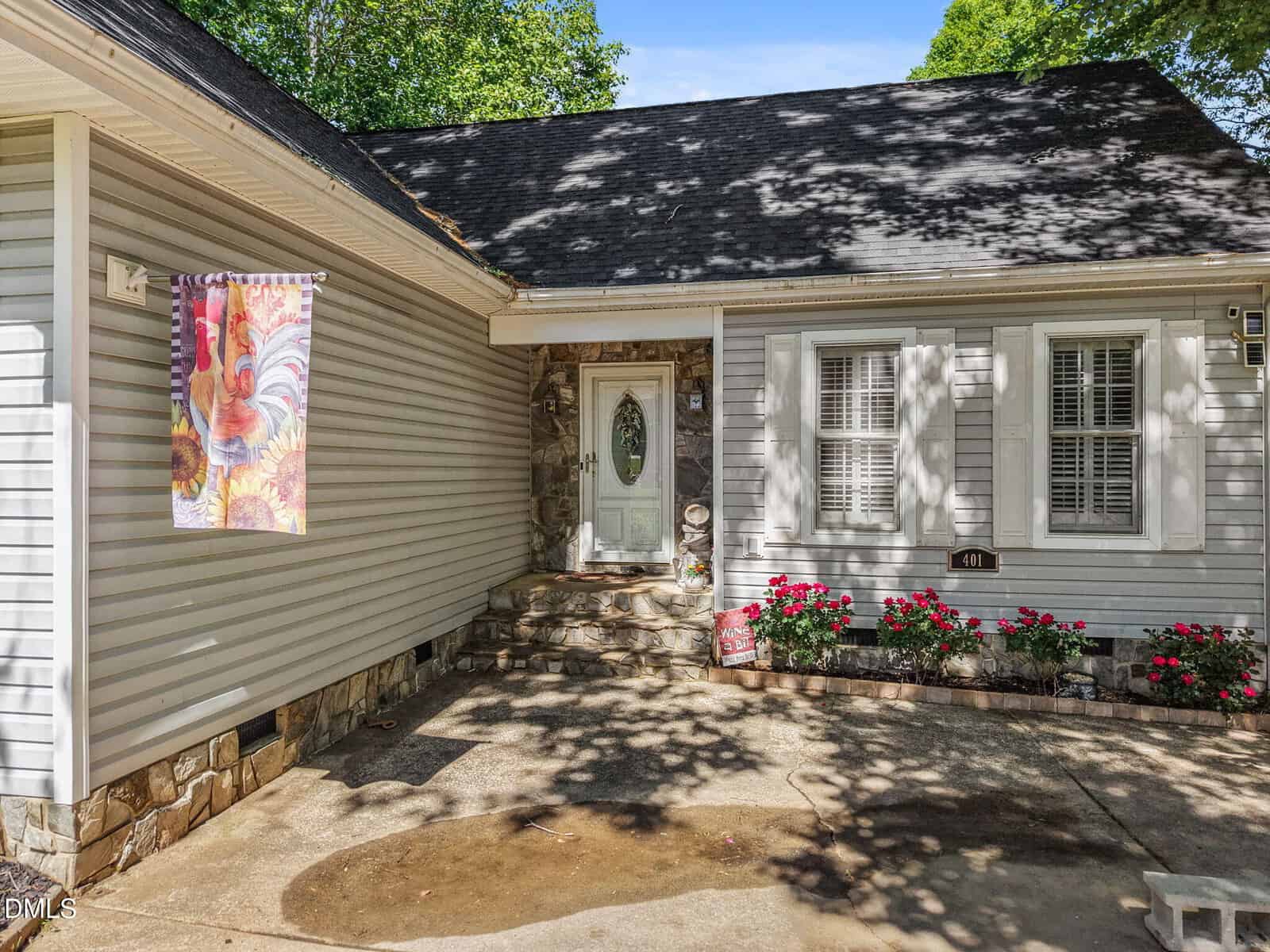 Single-story house with light gray siding, stone accents, and a white front door with an oval window. A colorful rooster flag hangs near the entrance, and red flowers are planted along the front wall. Trees cast shadows on the driveway.