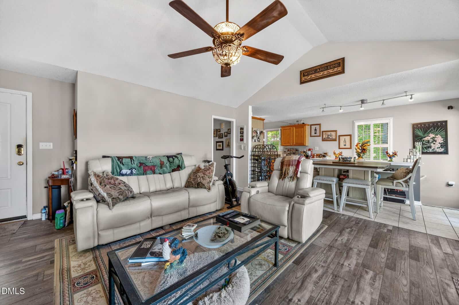 A cozy living room with light-colored sofas, a glass coffee table, and wood flooring. A ceiling fan hangs above. The space opens to a kitchen with bar seating and wooden cabinets. Decor and plants add warmth throughout.