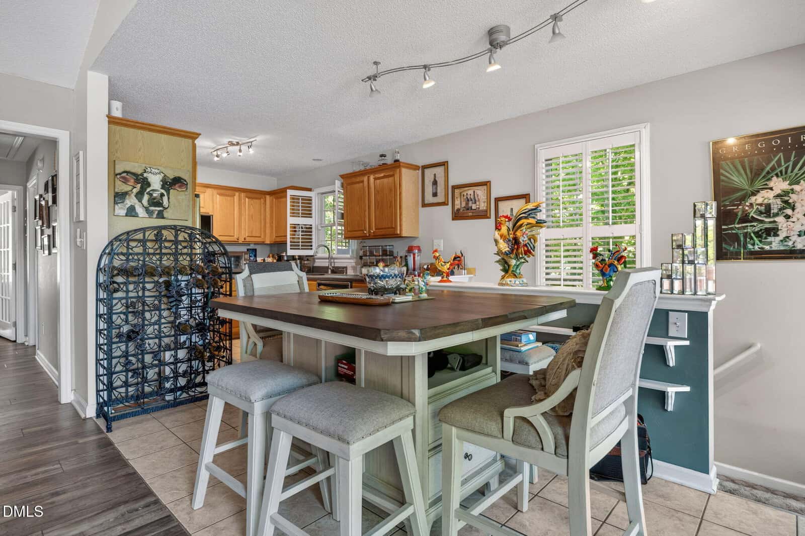 Bright kitchen and dining area with a sage green table, stools, wood cabinets, track lighting, plantation shutters, a metal wine rack, and colorful rooster decor.