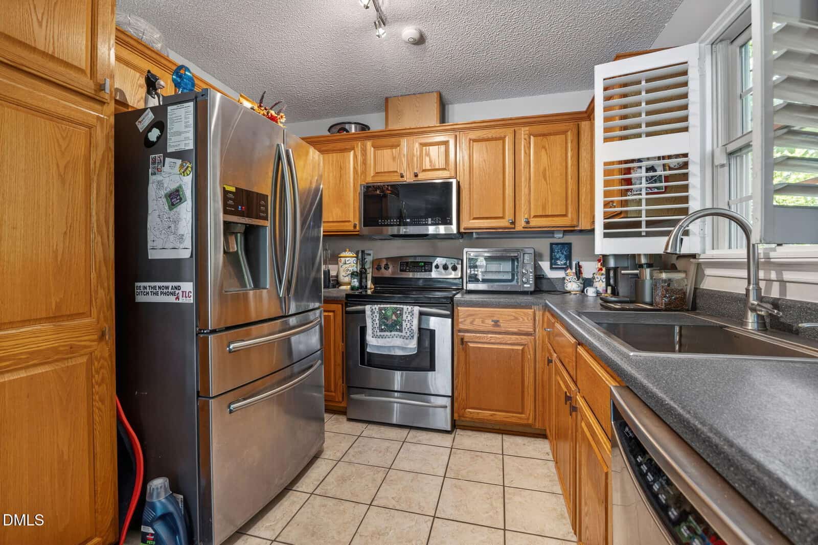 A kitchen with wooden cabinets, stainless steel appliances, a double-door fridge, a microwave, an oven, a dishwasher, a double sink under a window with white shutters, and tiled floors. Countertops hold small appliances and decor.