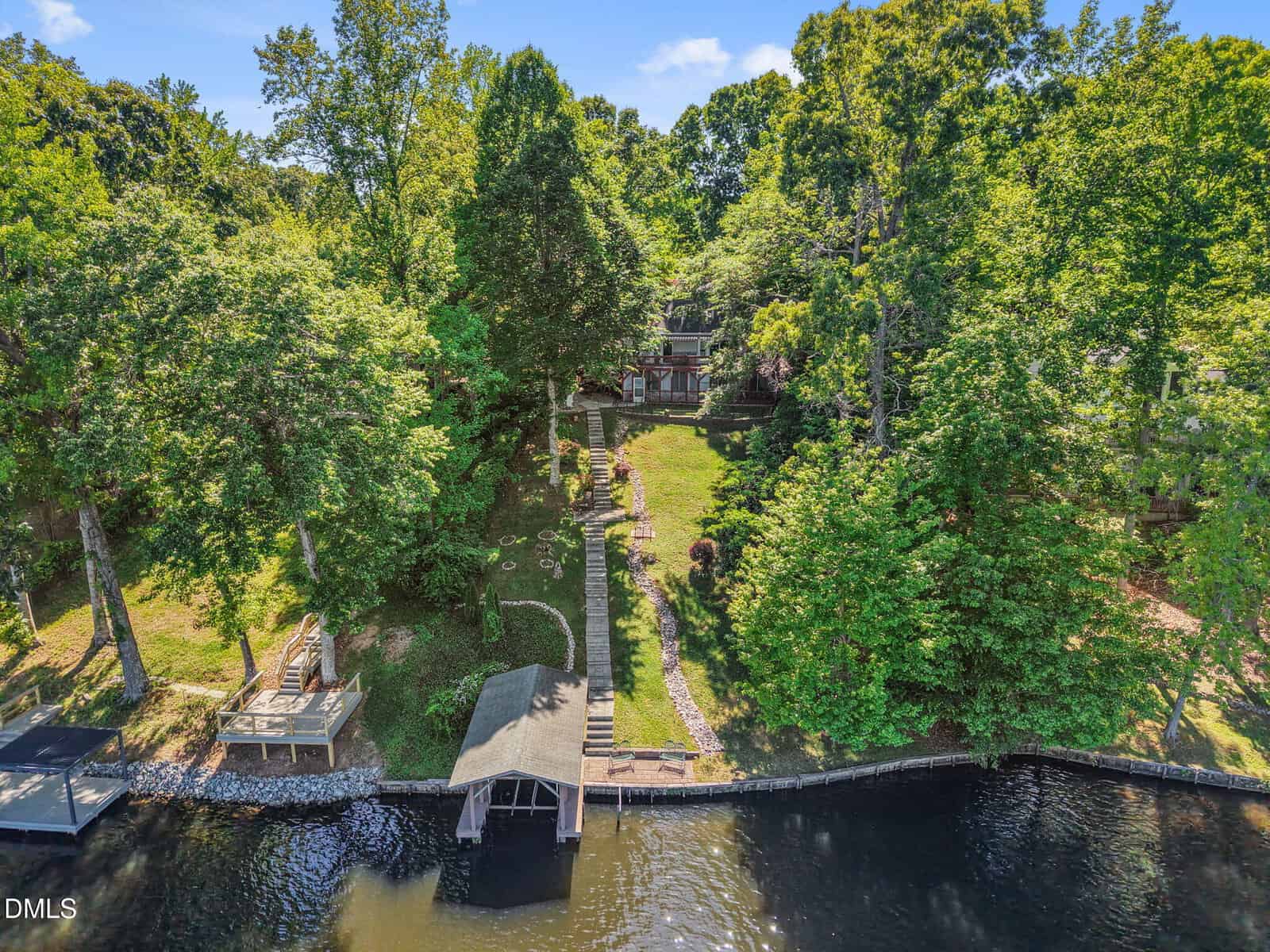 Aerial view of a lakeside property with a wooden dock and boathouse, lush green trees, terraced landscaping, and a house set back from the water among the trees.